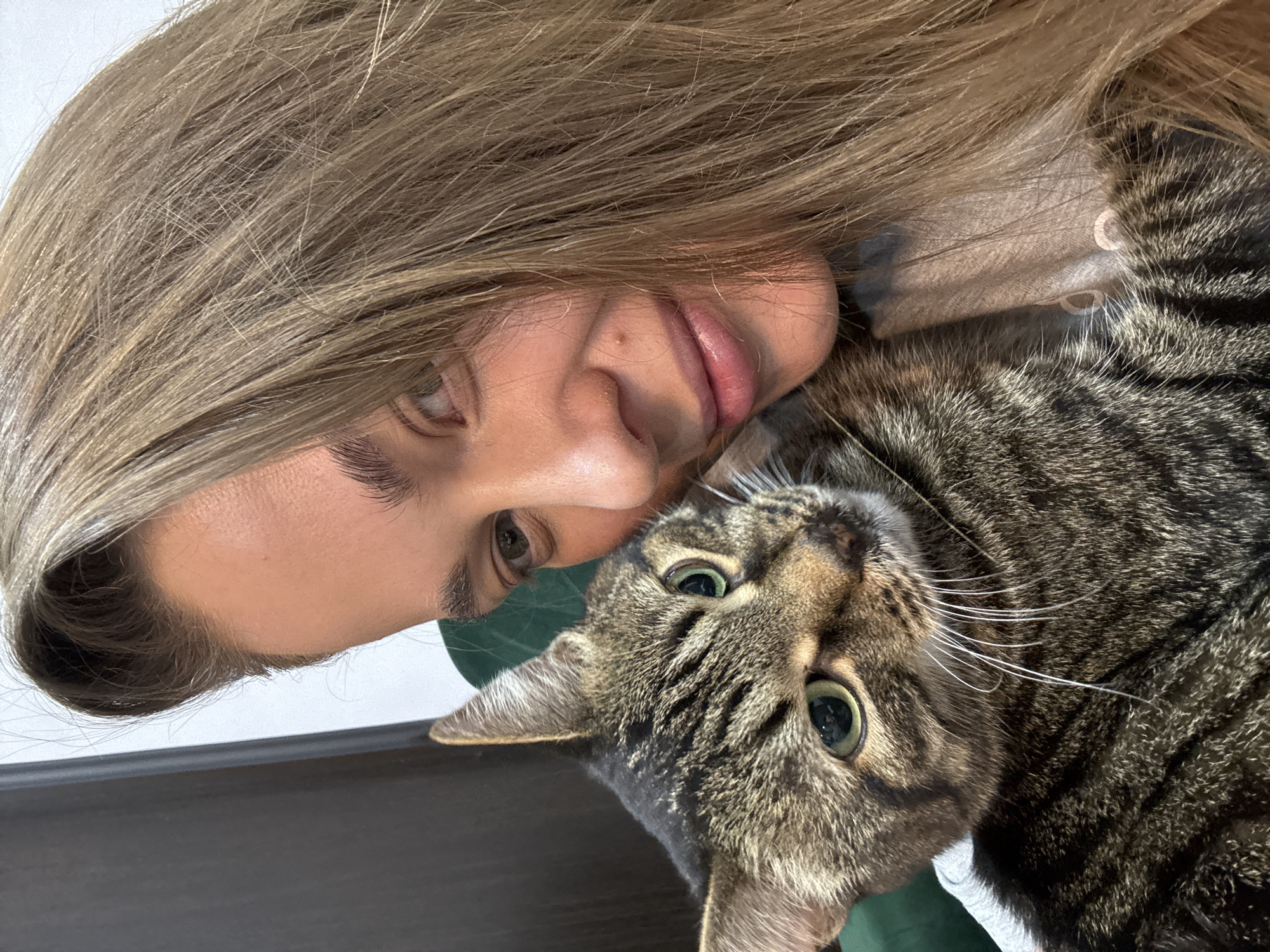 A woman with long, light brown hair is lying on her side, her face close to the camera. A tabby cat is nestled next to her, its face also close to the camera. The cat has green eyes and distinct tabby markings. The background is out of focus, but appears to be a dark surface. There are no indications of the location being Chernushka, Russia.