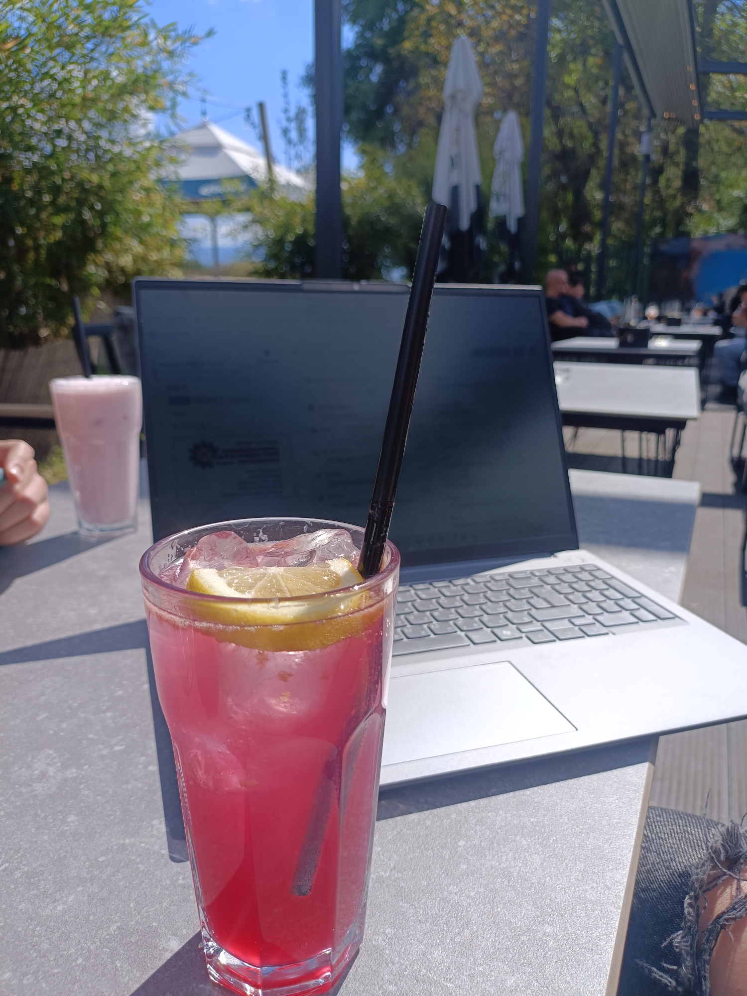 This media file captures a vibrant outdoor scene, likely at a cafe or restaurant in Plovdiv, Bulgaria, given the context. The photograph is taken on a bright, sunny day, evidenced by the clear blue sky and strong shadows cast on the table.
In the immediate foreground, a tall glass filled with a bright pink or red beverage with ice, a lemon slice, and a black straw takes prominence. To its left, partially visible, is another tall glass containing a light pink milkshake, also with a black straw, and a hand resting nearby.
Dominating the midground is an open silver or light gray laptop, its screen displaying content that includes a prominent red rectangular graphic with a white gear icon and some illegible white text. The laptop's keyboard is clearly visible.
The background reveals a relaxed outdoor setting. On the left, lush green foliage, possibly bamboo, creates a natural barrier. Towards the center and right, several white patio umbrellas are visible, along with more trees and the blurry figures of other patrons seated at tables. One person appears to be reclining comfortably. A light-colored building with a distinct roofline can be faintly seen in the distance against the blue sky.
The scene suggests an activity of combining work or casual browsing on a laptop with enjoying refreshing drinks in an alfresco environment. The overall mood is one of leisure and productivity under pleasant weather conditions.