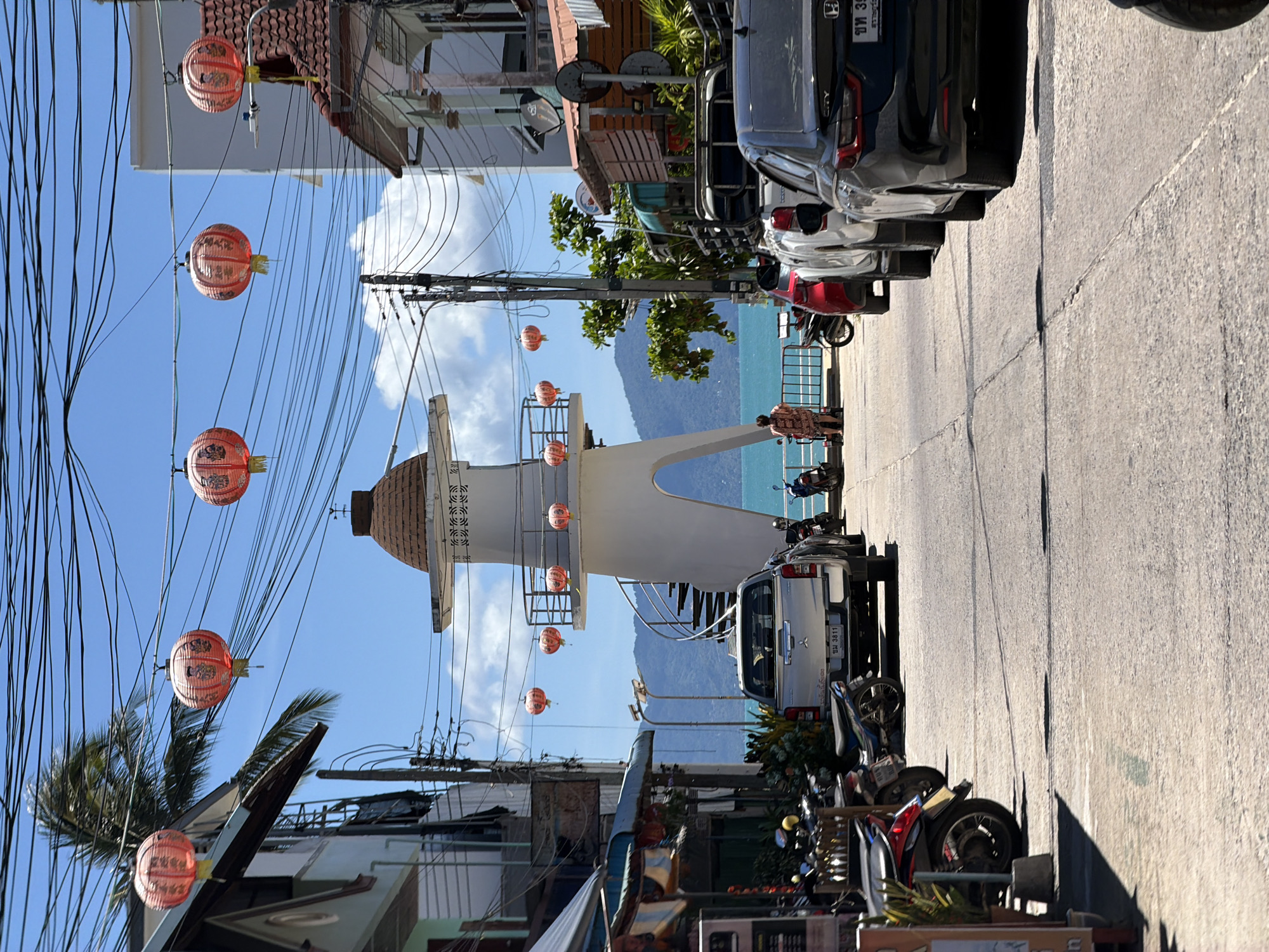 The image depicts a street scene in Baan Mae Nam, Thailand, characterized by a bright, sunny day with a clear blue sky. The foreground features a paved road lined with parked motorcycles and cars. On the left side of the road, there are buildings with awnings and signs, suggesting a commercial area. Further down the street, a white lighthouse-like structure with a spiral staircase is visible, overlooking the ocean. Red lanterns are strung across wires overhead, adding a decorative element to the scene. In the distance, a lush green mountain range meets the turquoise ocean. A woman in a patterned dress stands near the water's edge, facing the sea, while a few other individuals are present in the background. The overall atmosphere is one of a bustling seaside town, with a blend of local businesses and scenic natural beauty.