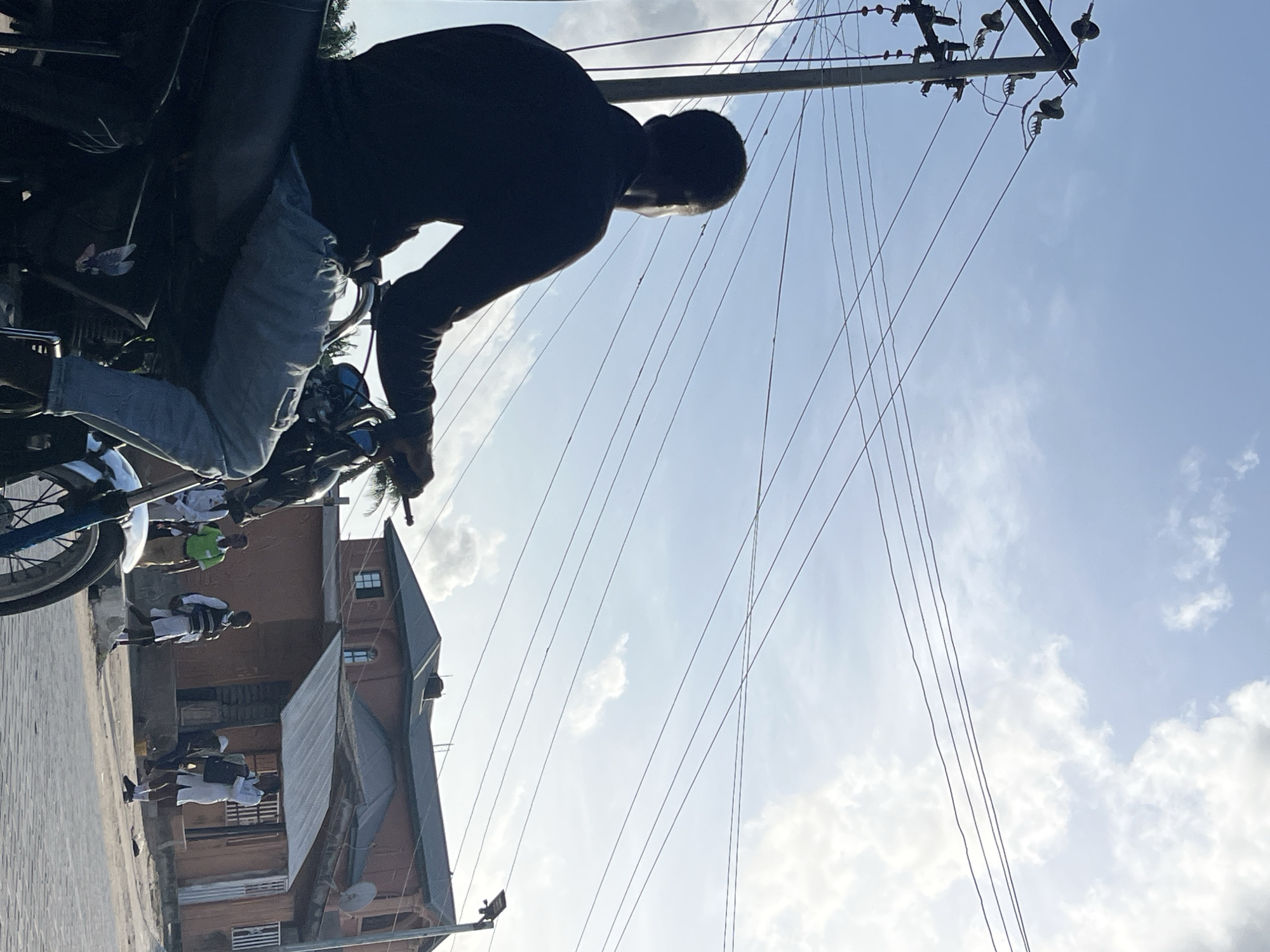 The image shows a man sitting on a motorcycle in Badagry, Nigeria. The scene also includes an electrical pole with numerous wires extending outwards. In the background, there's a two-story building with an adjacent satellite dish. Several people, presumably students in white and black uniforms, are walking along the street. The sky above is a light blue, filled with patchy clouds.