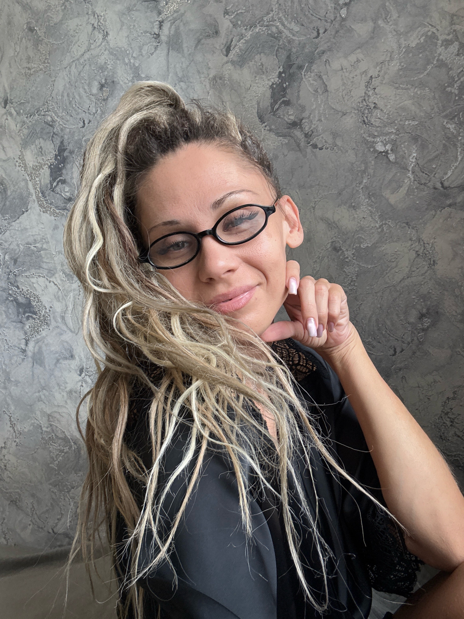 A medium shot shows a woman with blonde hair pulled into a high ponytail, wearing black-rimmed glasses and a black robe with lace detailing. She has her left hand resting on her chin, and she is looking at the camera with a slight smile. The background is a textured gray wall. There is no indication of the location being in Rusanovo, Russia.