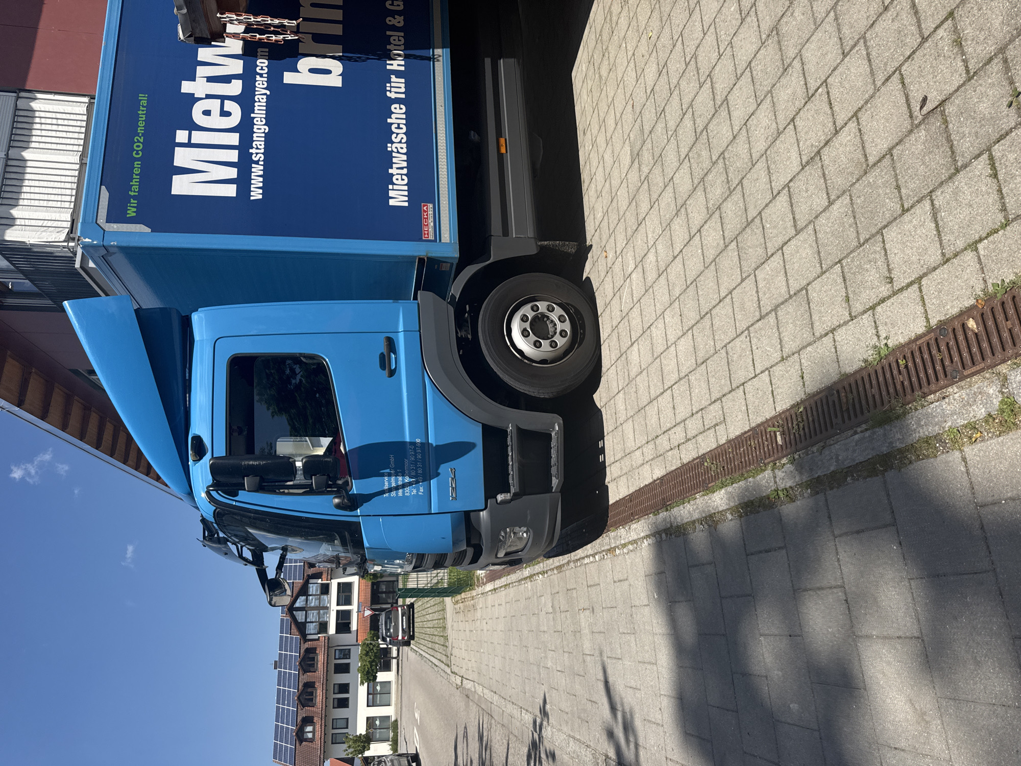 The image shows a blue commercial truck parked on a paved area next to a sidewalk. The truck has text on its side that reads "Mietwäsche für Hotel & G..." and "www.stangelmayer.com". The truck also has a sticker that says "HECKA". In the background, there are buildings with solar panels on their roofs, a street with parked cars, and a green fence. The sky is blue with some clouds. The scene appears to be in a residential or commercial area in Höhenkirchen-Siegertsbrunn, Germany. The truck is the primary subject, and it is stationary. The environment is sunny and appears to be a daytime scene.
