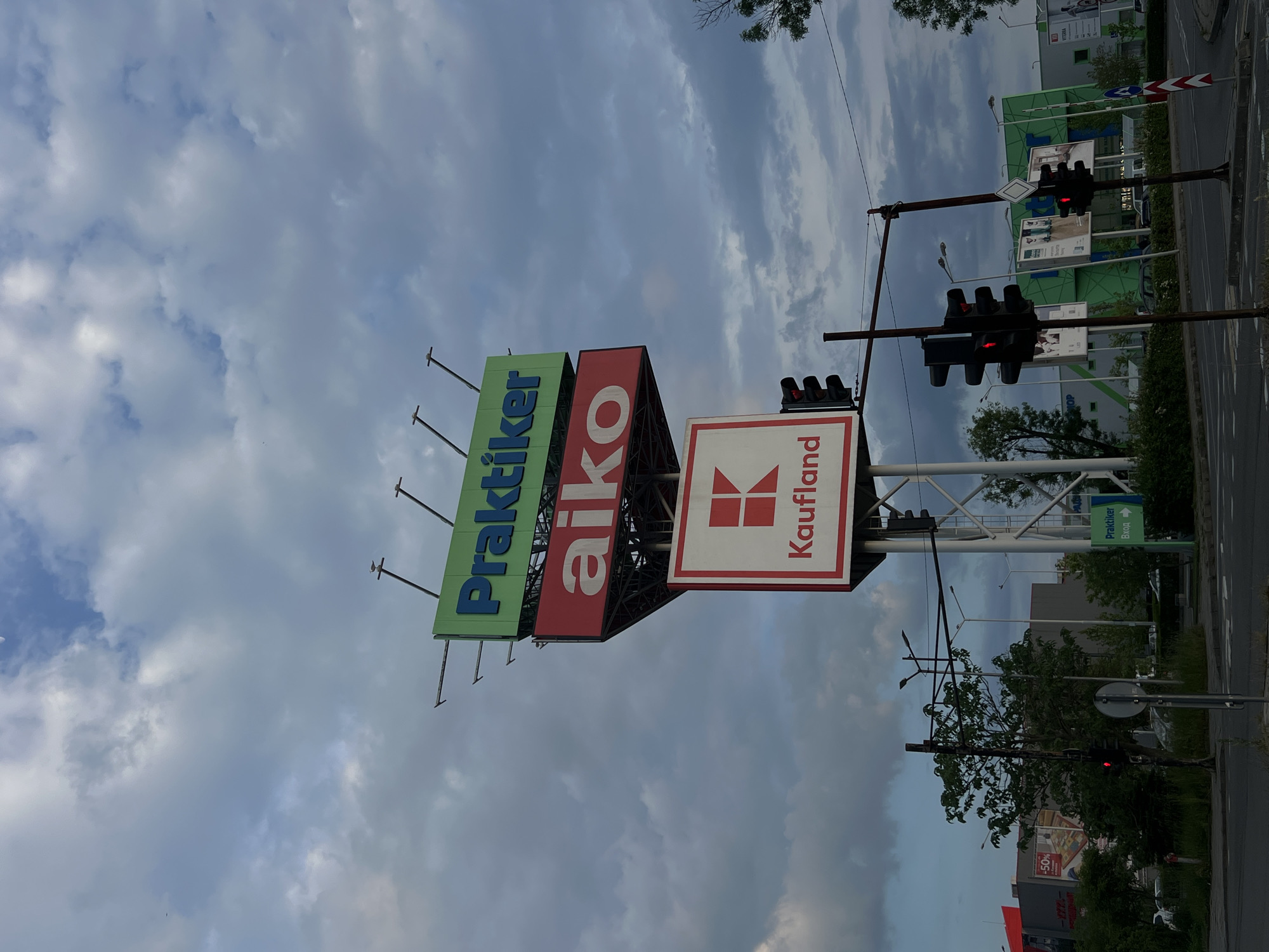 The image captures a vertical view of a commercial area, likely in Burgas, Bulgaria, based on the signage visible. The primary focal point is a multi-branded sign tower featuring the logos of "Praktiker," "aiko," and "Kaufland," all prominently displayed. The sky behind the sign tower is overcast with grey and white clouds.
In the background, commercial buildings, traffic lights showing red, streetlights, and greenery are visible. A road runs along the right side of the frame. The scene appears to be a busy commercial area at dusk.