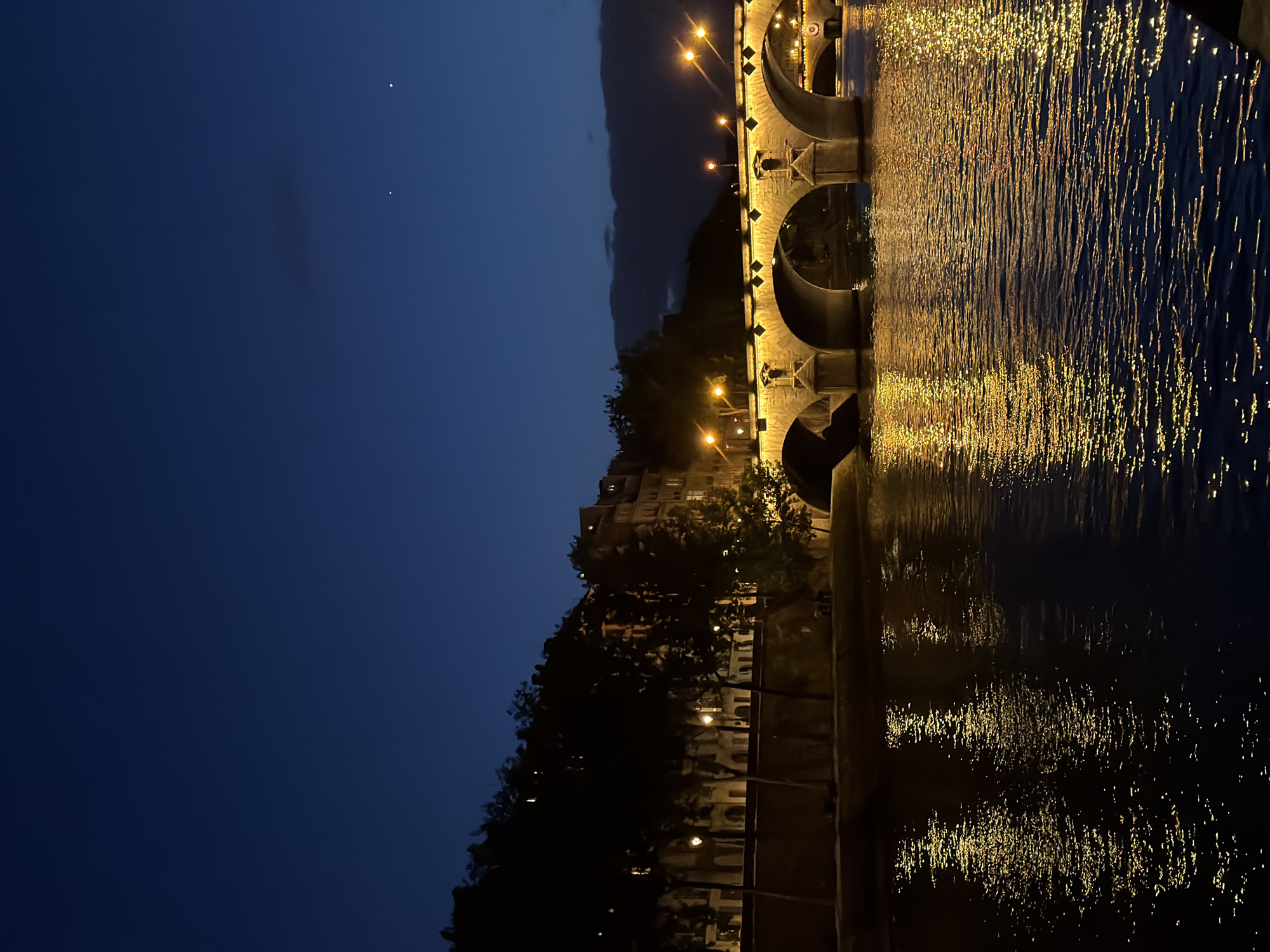 The image depicts a nighttime scene of the Pont Neuf bridge spanning the River Seine in Paris, France. The sky is a deep, dark blue, indicating twilight or night, with faint stars visible in the upper right quadrant and a band of dark clouds low on the right horizon.
The River Seine occupies the foreground and midground, its dark surface reflecting the numerous artificial lights with shimmering golden-yellow patterns caused by ripples. The **Pont Neuf**, a historic stone arch bridge, is the central subject, brightly illuminated by warm yellow lights that accentuate its architectural details, including its multiple arches, robust stone piers, and distinctive diamond-shaped bossages above the arches. Small, semi-circular structures (niches/boutiques) are visible on the bridge's piers. Beyond the far end of the Pont Neuf, a portion of a second, smaller, also lit bridge can be seen.
Along the left bank (Rive Gauche), a dense line of dark trees partially obscures multi-story buildings, from which warm light emanates from windows. Several streetlights line a riverside promenade, casting additional warm glows. Small, indistinct human figures are discernible on this promenade. Buildings on the far bank (Rive Droite), beyond the bridge, are also visible, showing warm internal lighting. The entire scene is bathed in the warm, golden glow of artificial illumination, creating a stark contrast with the dark sky and river.