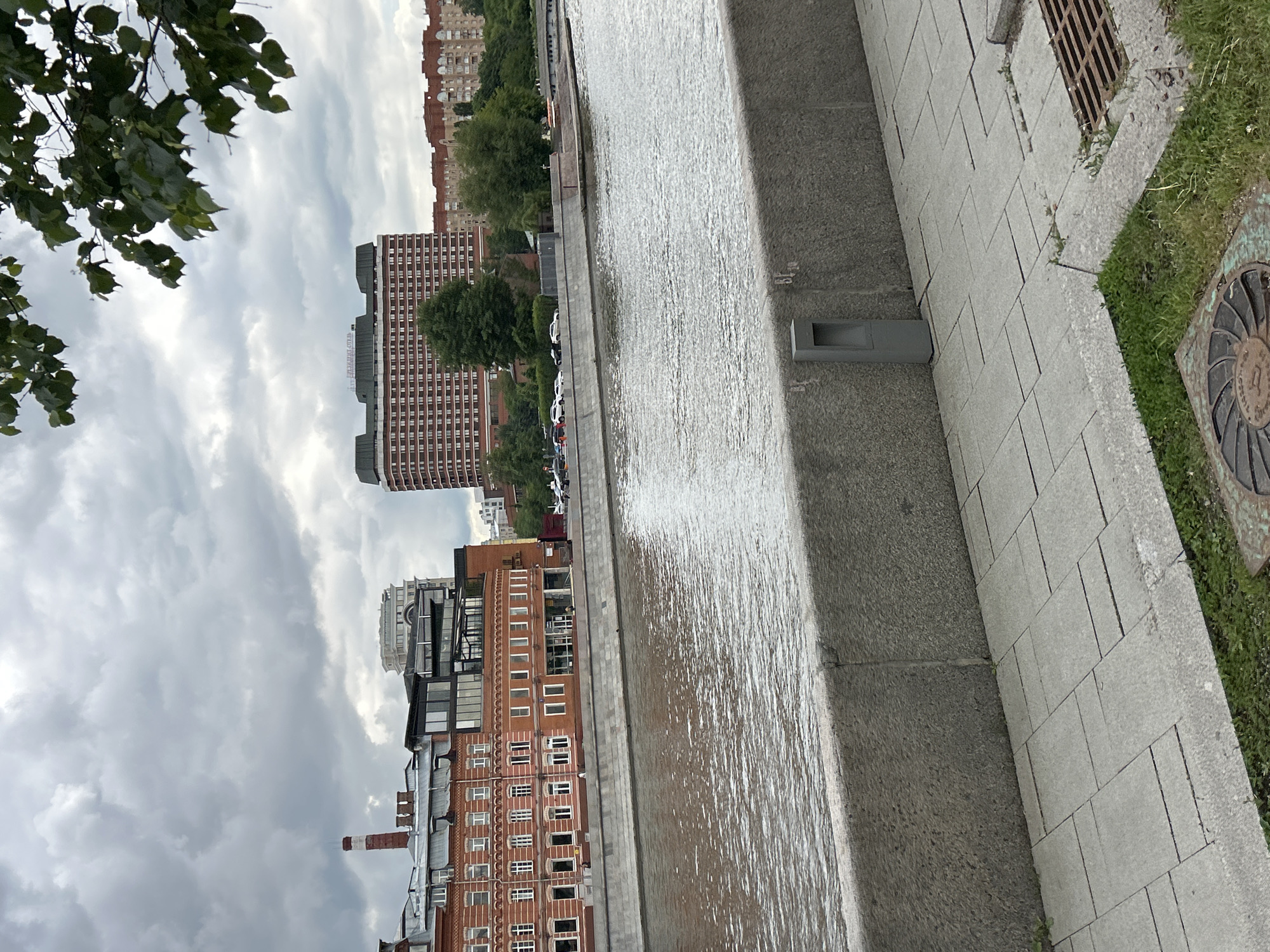 The image shows a view along a riverbank in an urban setting, possibly a city. On the left, there is a series of buildings including brick structures with windows and other modern buildings. In the middle, there is a body of water with a stone edge and light reflecting off the surface. On the right, there is a paved area with stone tiles, some patches of grass, a drain, and a circular metal object (possibly a manhole cover) on the ground. The sky is cloudy with an overcast appearance. Some tree branches are visible in the top left corner.