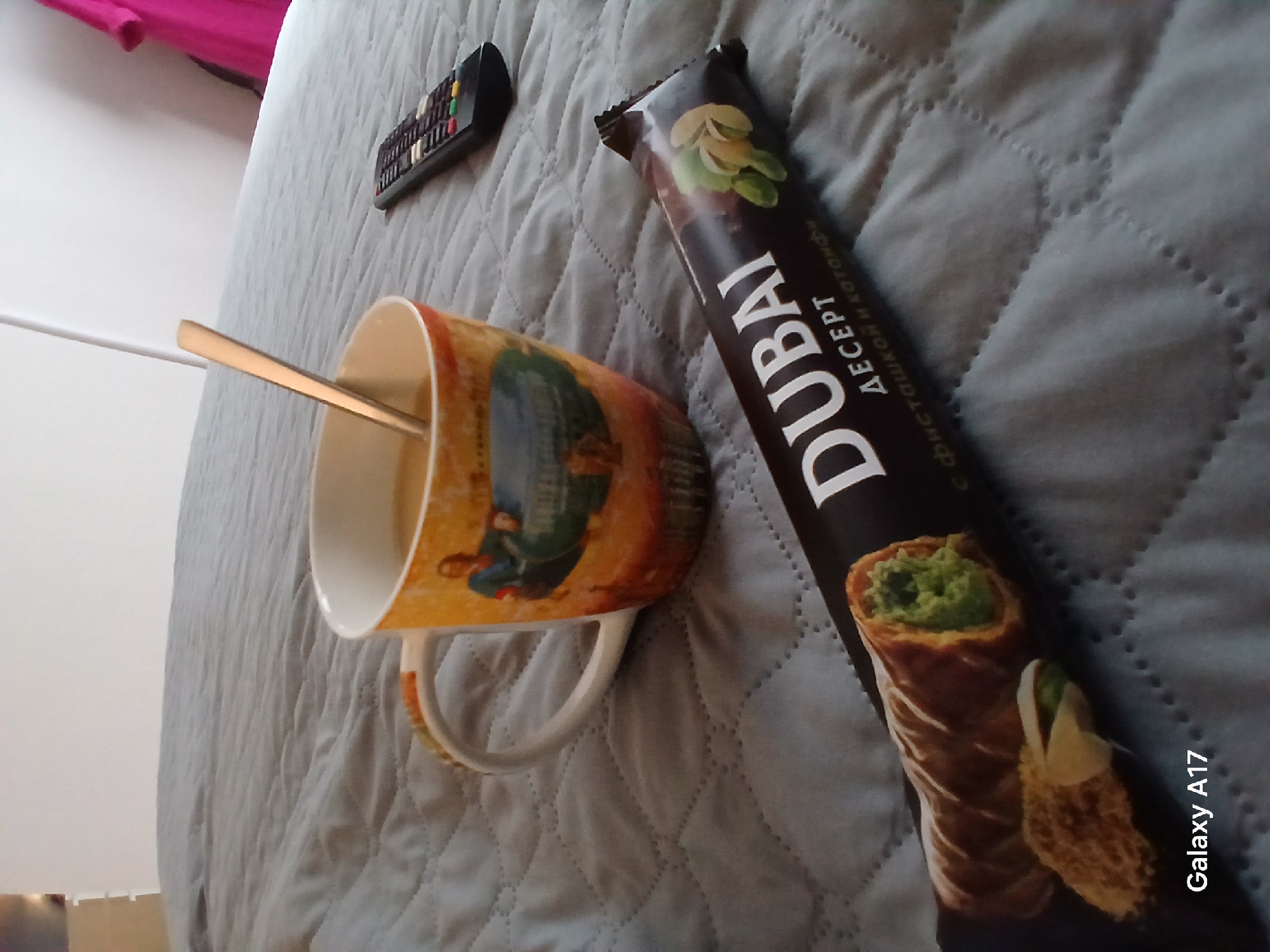 The image shows a close-up shot on a quilted grey surface, likely a bedspread. A mug with a spoon inside it sits near a black candy bar labeled "DUBAI" with pistachios pictured on the packaging. A remote control is visible further up on the surface. The presence of Russian Cyrillic text on the candy bar packaging suggests the image may have been taken in Russia, but there are no details pointing to the specific city of Yekaterinburg. The mug is not unique and could be found anywhere. There are no people visible in the image.