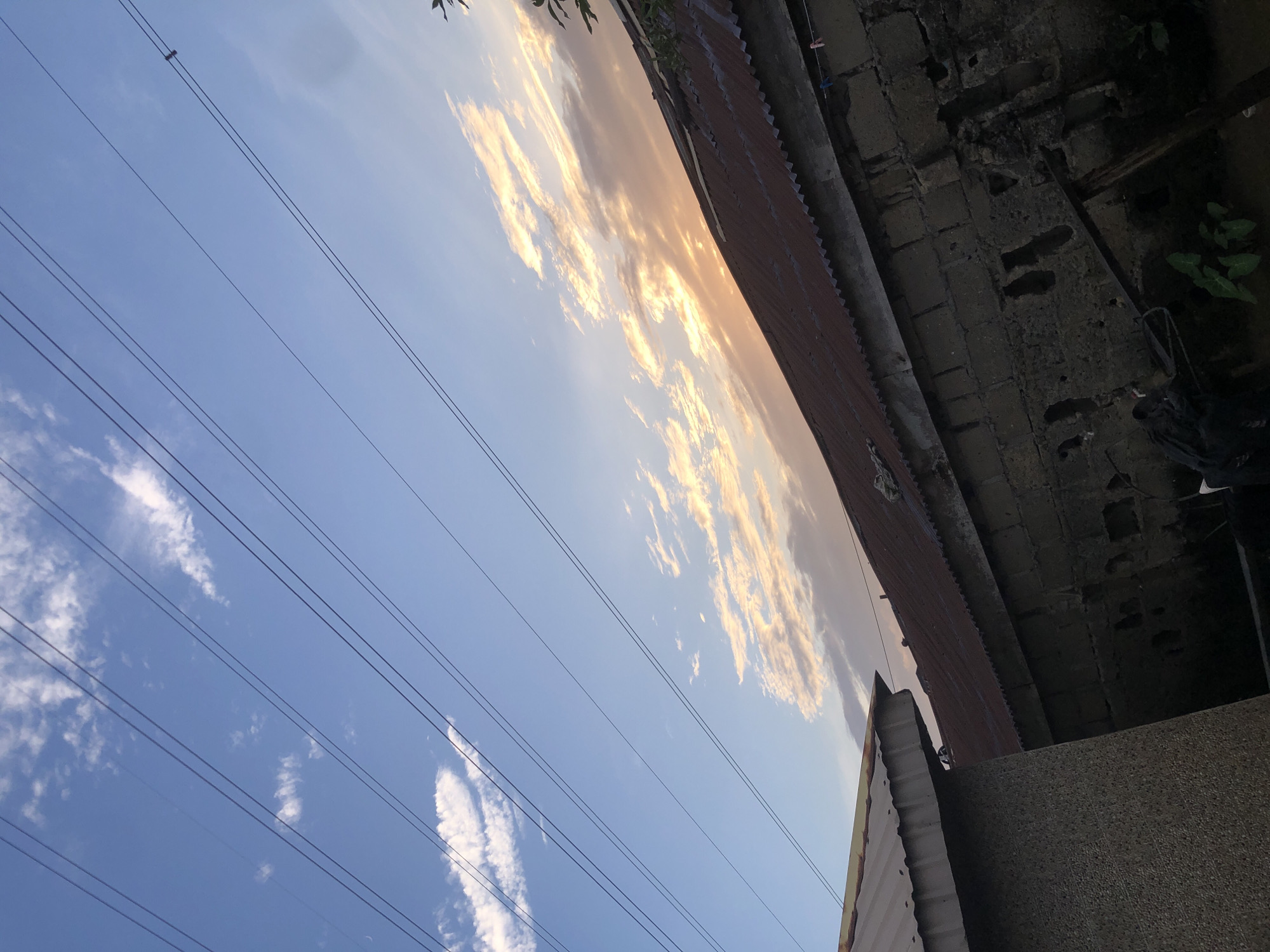 The image depicts a view of the sky and rooftops in Ado, Nigeria, during what appears to be dusk. The sky is a gradient of blue, transitioning to a warm orange and yellow where the sun is setting, illuminating scattered clouds. Several parallel power lines stretch across the upper portion of the frame. In the foreground, the corrugated iron roof of a building is visible, angled downwards. Below the roof, a textured brick wall forms part of a structure, with some dark foliage growing at its base. The scene suggests an outdoor, urban or semi-urban environment at the end of the day.