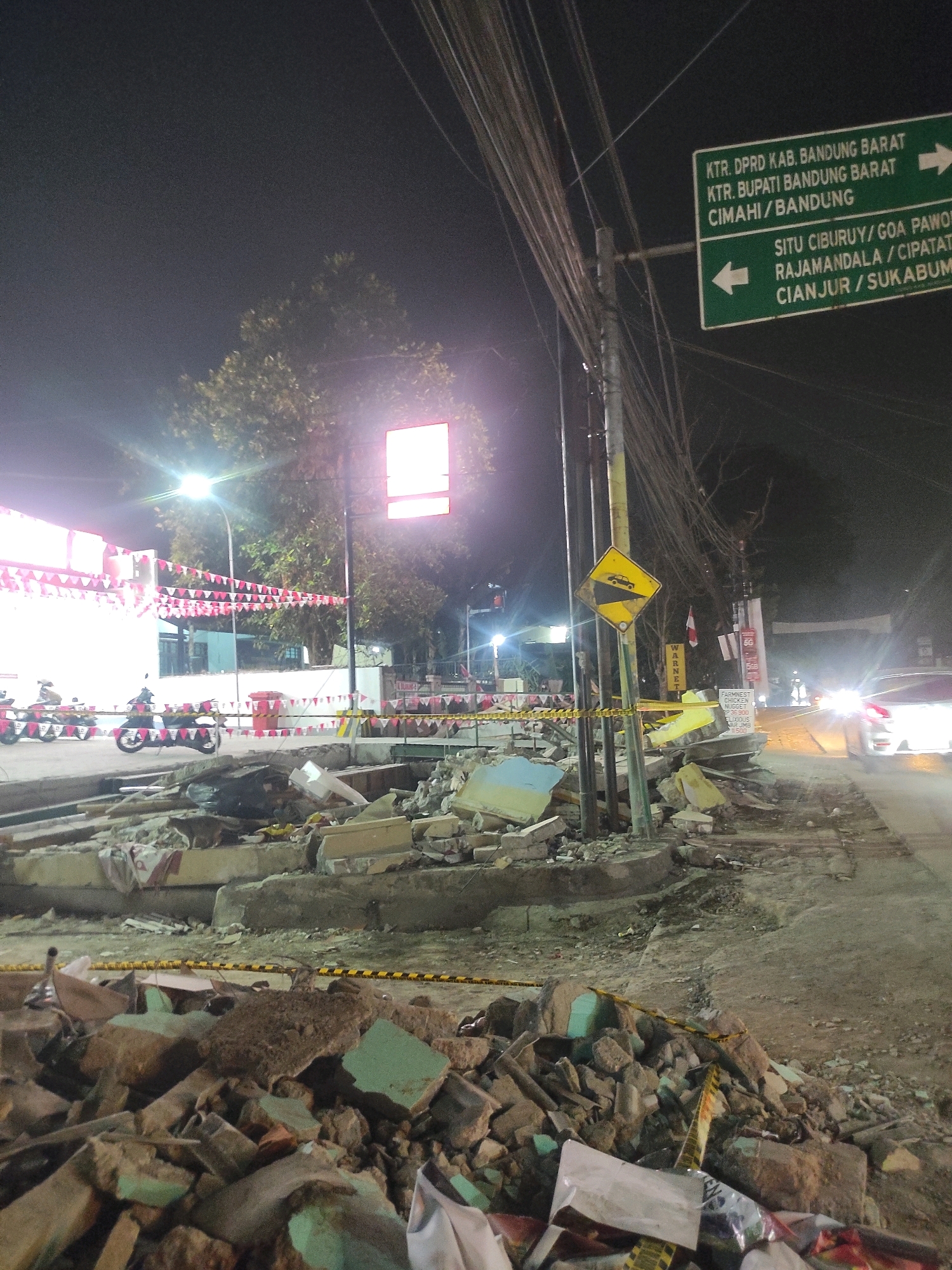The image depicts a street scene at night in Padalarang, Indonesia, following what appears to be a demolition or a structural collapse. In the foreground, a large pile of rubble, consisting of broken concrete, bricks, and debris, is visible. Yellow and black caution tape is draped across the rubble and the surrounding area, indicating a hazardous zone. Further back, a road is visible with caution tape lining its edge. Several motorcycles are parked on the side of the road. A building with white walls and red and white pennant banners strung across it is also present. In the background, trees and a lit sign can be seen, along with streetlights illuminating the scene. A car with its headlights on is approaching from the right side of the image. Road signs are mounted on a pole, one of which is a yellow diamond-shaped sign depicting a car descending a slope, indicating a steep downgrade. Another sign is a green rectangular sign with white lettering providing directional information to various locations. The overall atmosphere suggests a post-event scenario, possibly related to construction, destruction, or an accident.
