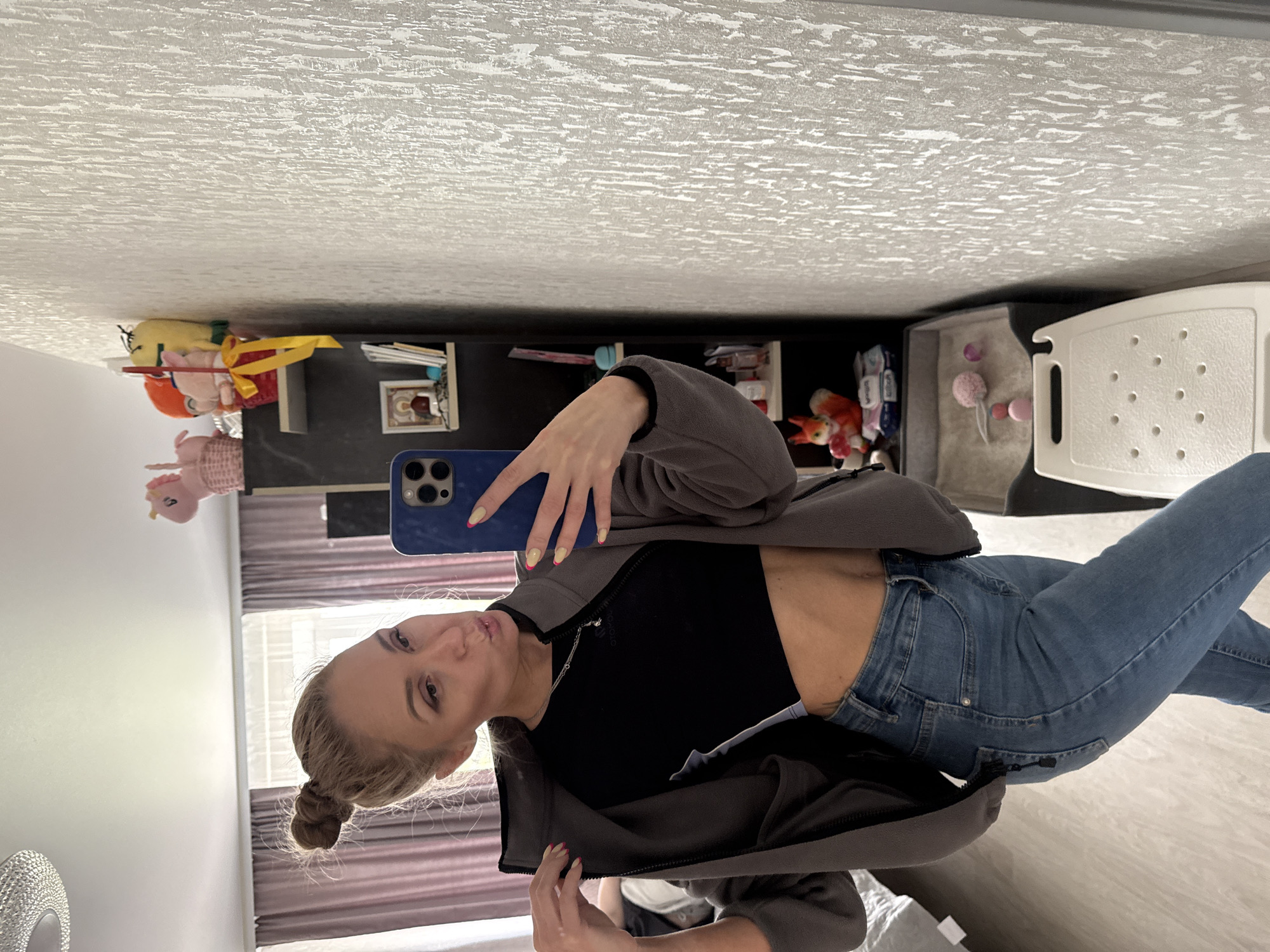 A young woman is taking a selfie indoors. She is wearing a black crop top, blue jeans, and a gray fleece jacket. Her hair is blonde and styled in a bun. She is holding a blue smartphone to take the picture. In the background, there is a shelf with toys and other items, a window with curtains, and a white chair. The location is likely a home in Vereshchagino, Russia.