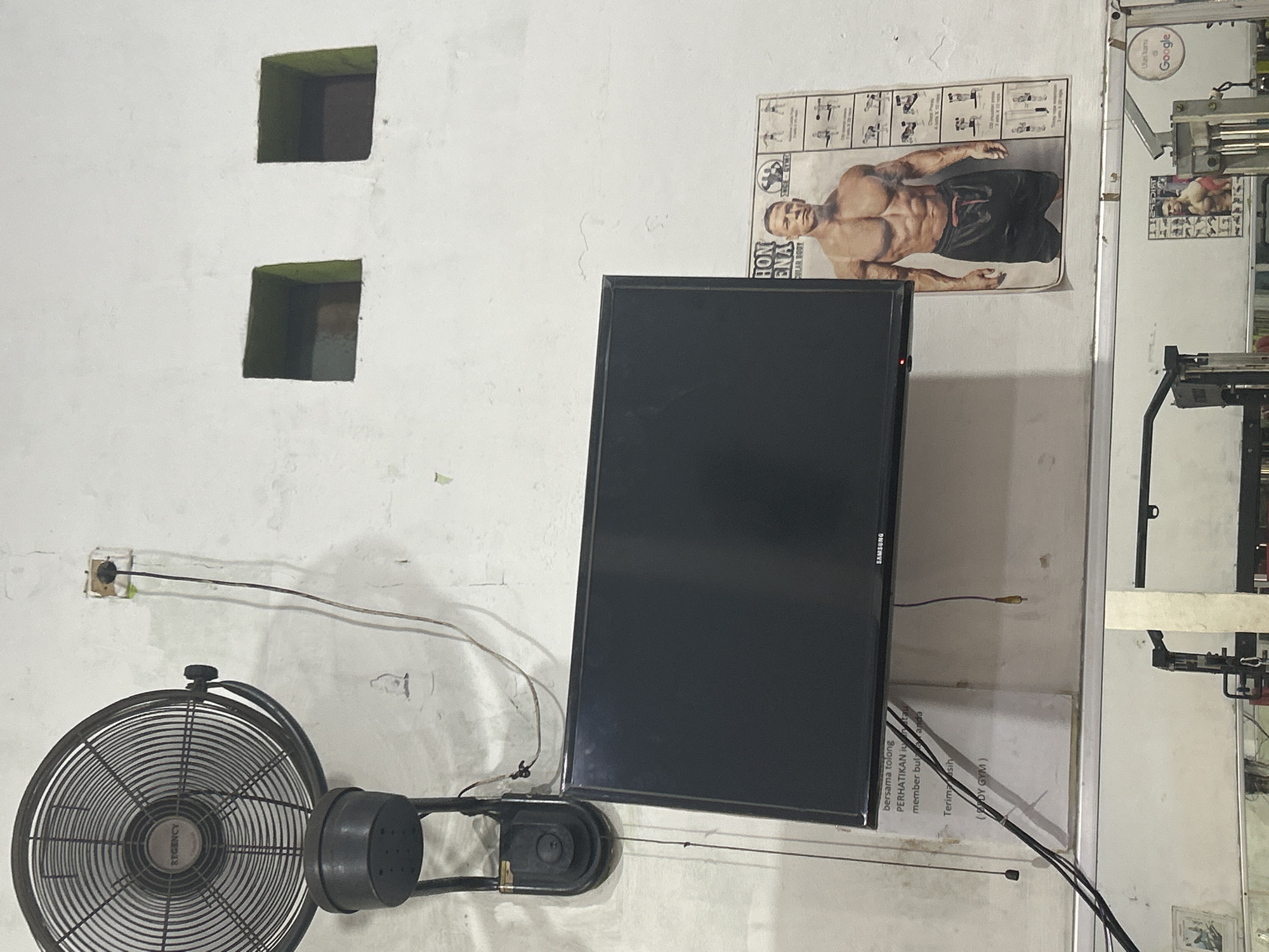 The image depicts an interior scene, likely a gym in North Jakarta, Indonesia, as indicated by the prominent Google logo reflected in a mirror and the presence of fitness equipment. A large, industrial-style fan is mounted on the wall in the upper left corner. Below it, a black flat-screen television is mounted on the wall. To the right of the TV, a poster featuring a muscular man, likely a bodybuilder or athlete, is displayed. The walls are painted white and appear somewhat weathered, with visible cracks and imperfections. Two small, dark square openings are present in the wall above the TV. Electrical wires and a power outlet are visible near the fan and TV. In the foreground, a mirror reflects gym equipment and another poster, also featuring a muscular man. A Google logo is visible on a sign in the reflection. The overall environment suggests a functional, no-frills gym setting.