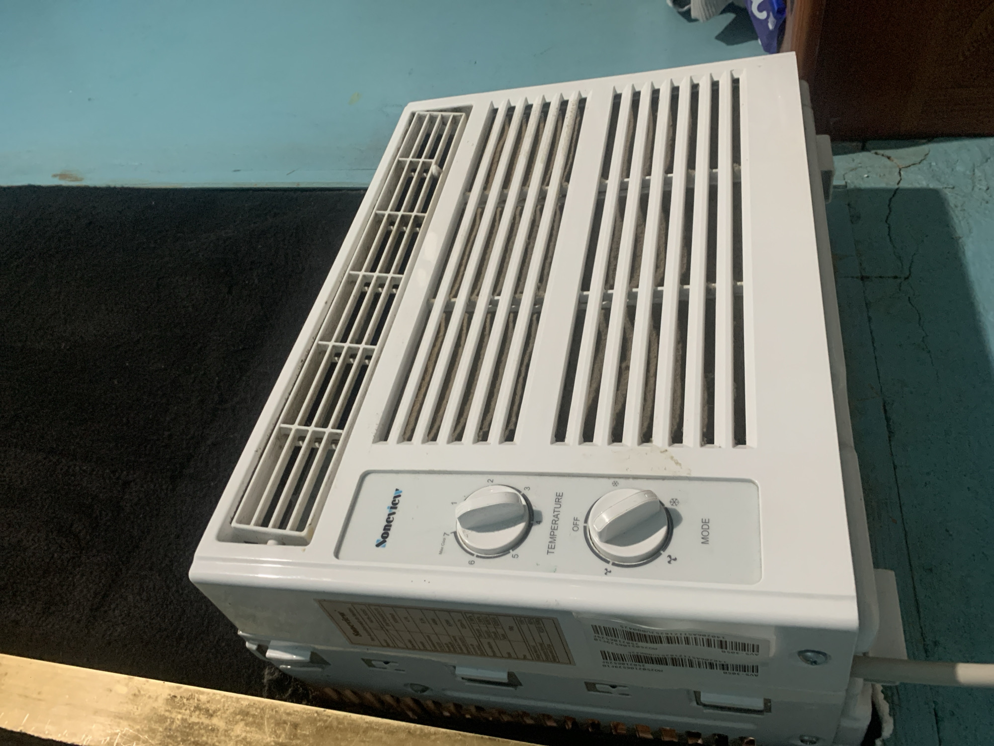 A white window air conditioner sits atop an unknown object. The air conditioner is oriented at an angle with the front panel and control knobs visible. The model appears to be a "Soneview" unit, with knobs for temperature and mode settings. The unit has visible slats and vents for airflow. The air conditioner rests on a surface that is partially visible at the bottom, with dark carpet and a light blue wall in the background. Part of a brown piece of furniture is visible behind the air conditioner. The setting appears to be indoors. There are no people visible. There is no information in the image to indicate it is in Santa Ana, Venezuela.