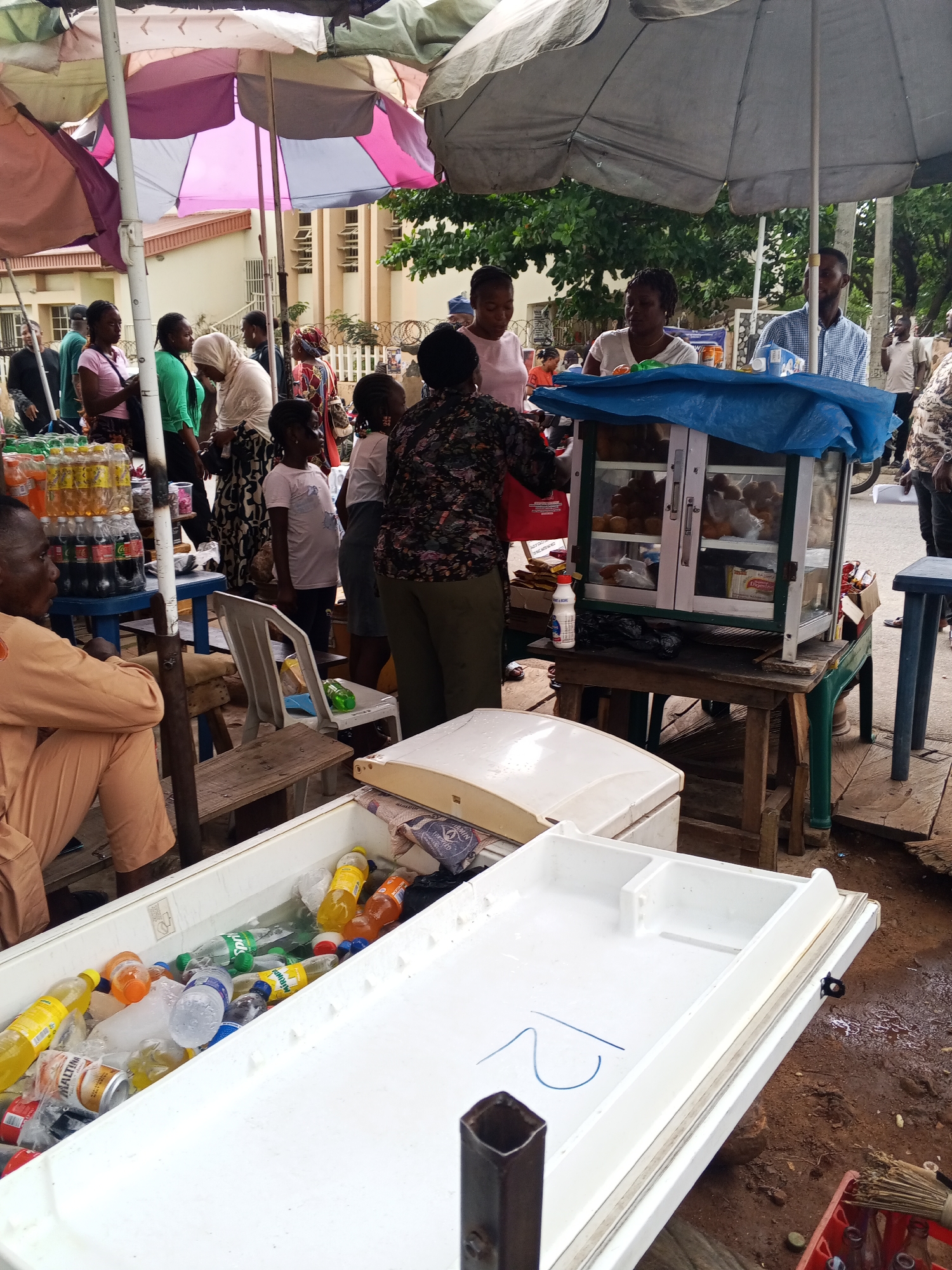 In Lokoja, Nigeria, an outdoor market scene unfolds. Several individuals are gathered under colorful umbrellas and a large gray tarp. In the foreground, a white cooler filled with various bottled drinks, including what appear to be sodas and water, rests on the ground. A man in a tan outfit is seated to the left of the cooler, looking away from the camera. Behind him, a white chair sits near a wooden stand. Further back, a glass display case filled with food items is set up on a wooden table. A woman in a patterned shirt is attending to customers at this stand. Multiple people, including women and children, are visible interacting and standing around the market stalls. The ground is dirt, and the surrounding environment suggests a typical urban street market.