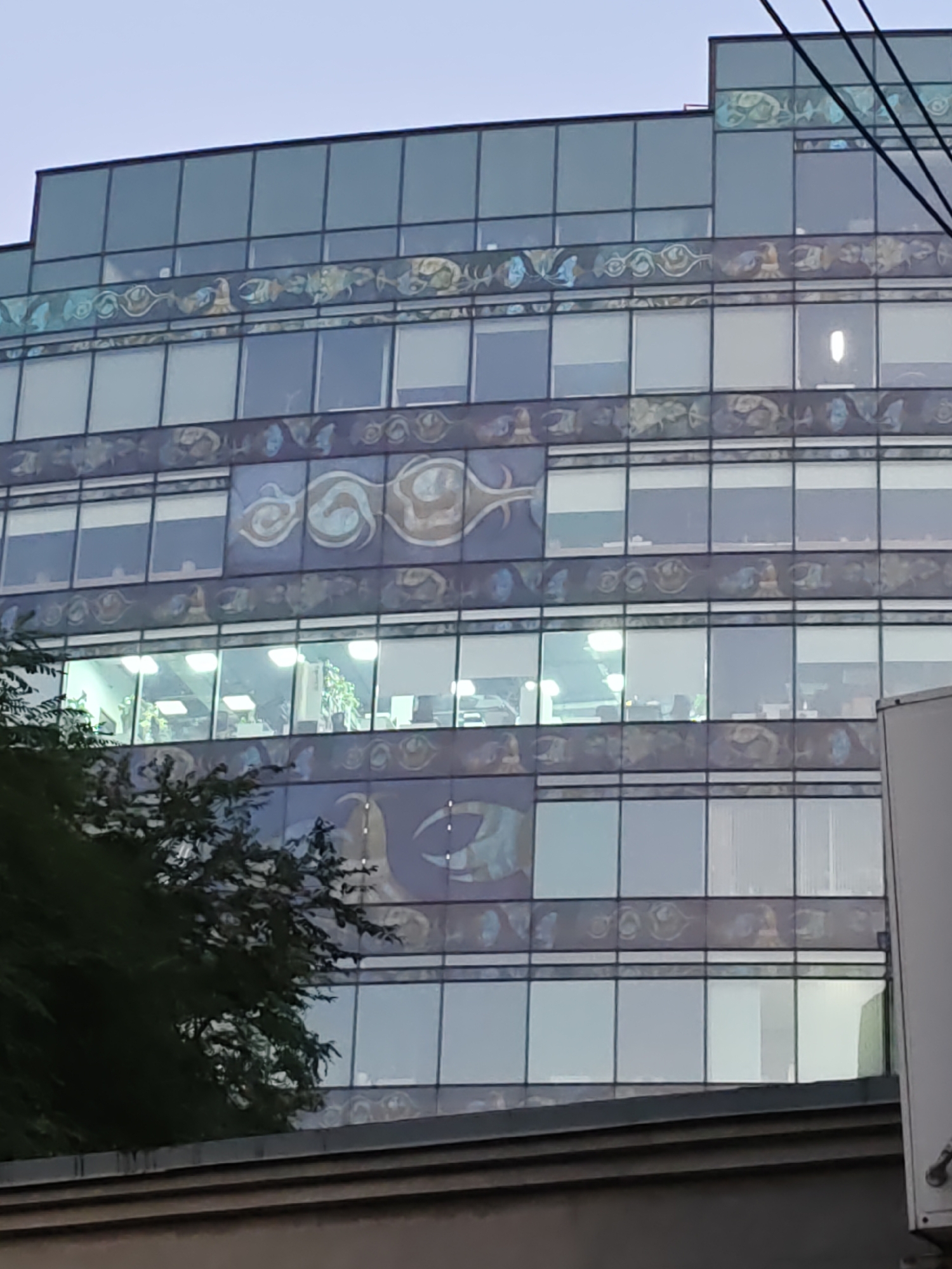 A modern office building in Rostov-on-Don, Russia, features a distinctive facade with alternating bands of windows and decorative panels. The decorative panels are adorned with intricate, abstract patterns in shades of blue, gold, and white, resembling stylized aquatic life or flowing organic forms. Some of the windows on the lower floors are illuminated, revealing the interior of an office space with desks, chairs, and potted plants, suggesting it is either a late evening or the building is occupied by businesses with extended hours. The building's architecture is contemporary, with a glass and metal exterior. The overall scene is dominated by the building's structure, with a glimpse of green foliage from a tree on the left, partially obscuring the lower levels of the structure. The sky above is overcast and grey.