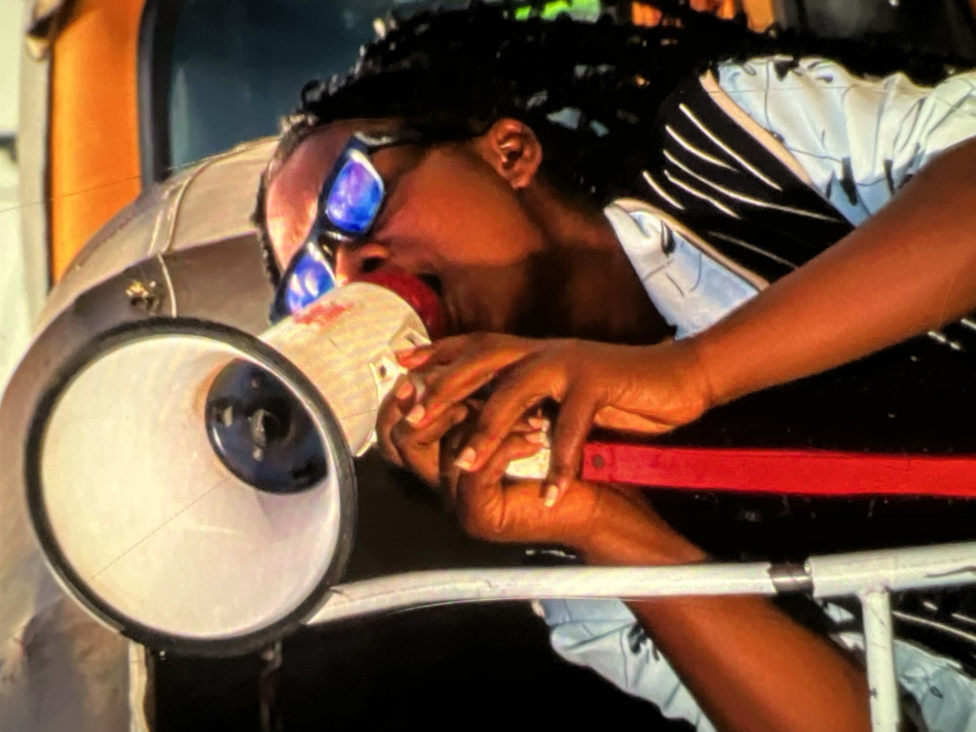 In Lagos, Nigeria, a person wearing sunglasses and a black vest over a light blue shirt with a patterned design holds a red and white megaphone to their mouth, appearing to speak into it. The megaphone has a black speaker grill visible inside the white horn. The person has dark skin and long, dark dreadlocks. Behind them, an orange vehicle is visible, suggesting an outdoor urban setting. The image is cropped tightly, focusing on the person and the megaphone.