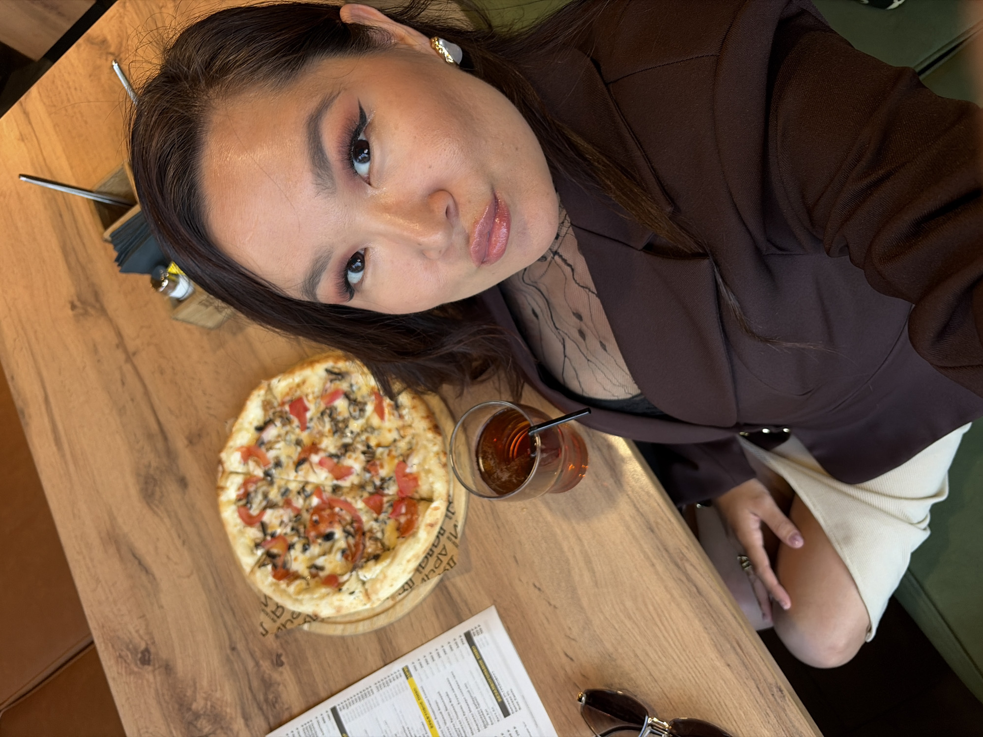 An angled selfie captures a woman sitting at a wooden table in a cafe or restaurant in Izhevsk, Russia. The woman, who appears to be of East Asian or Southeast Asian descent, looks directly at the camera with a slight pout. She has dark hair, styled with prominent winged eyeliner, pinkish eyeshadow, and glossy lips. She wears a dark brown blazer over a sheer black top with a dotted pattern, and a light beige ribbed skirt. A gold and white ear cuff adorns her right ear.
On the wooden table in front of her is a personal-sized pizza, topped with mushrooms and diced tomatoes, served on a round wooden board with patterned paper underneath. Next to the pizza, an amber-colored drink with a black straw sits in a clear glass. Other items visible on the table include a white menu with small print and yellow highlighted sections, a pair of black sunglasses with gold accents, and a metal holder containing napkins and cutlery. The background shows hints of green and brown upholstered seating, suggesting a cozy dining environment. Text partially visible on the pizza paper includes "S A H A" and "D A P U N U", likely part of a restaurant or brand name.