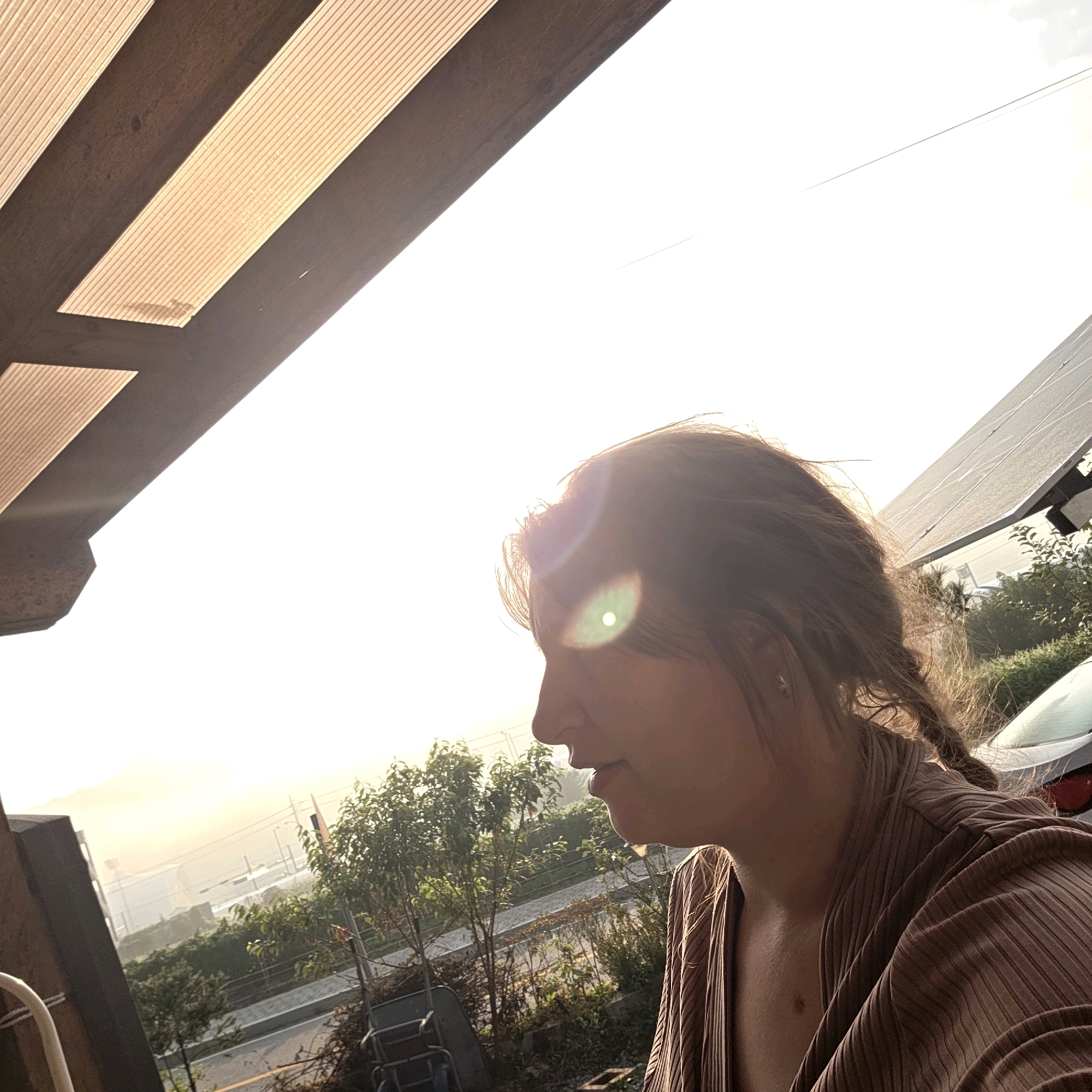A young woman with brown hair in braids is pictured from the side, looking out at a bright sky, possibly at sunset. She is wearing a brown, ribbed top. The background features solar panels on a roof, greenery, and a distant view of a town or city with buildings and a body of water. A car is partially visible on the right side of the frame. The foreground includes architectural elements resembling a covered patio. The overall scene is bathed in the warm light of the setting sun. The location is Dunnae-myeon, South Korea.