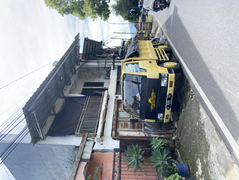 The image shows a yellow truck parked on the side of a paved road in the village of Rancabali, Indonesia. The truck is a dump truck with a black tarp covering the bed. The truck has a "Bismillah" sticker on the windshield and a sticker on the hood that reads "Permanana" with a crown logo. The truck has a license plate that reads "D 8389 YA". The truck is parked on the edge of a curb, with overgrown greenery next to it. The road curves to the right, with a single motorcycle riding towards the camera. The road leads to a residential area with houses and buildings on the left. The houses have flat roofs and are painted white, with some having pink or blue accents. There are power lines running above the road. The weather is overcast, with a gray sky and some clouds visible. The houses have railings and balconies made of metal. There are visible cracks in the walls of the buildings. The street is paved with asphalt and has a white stripe marking the edge. There is a concrete curb along the edge of the road.