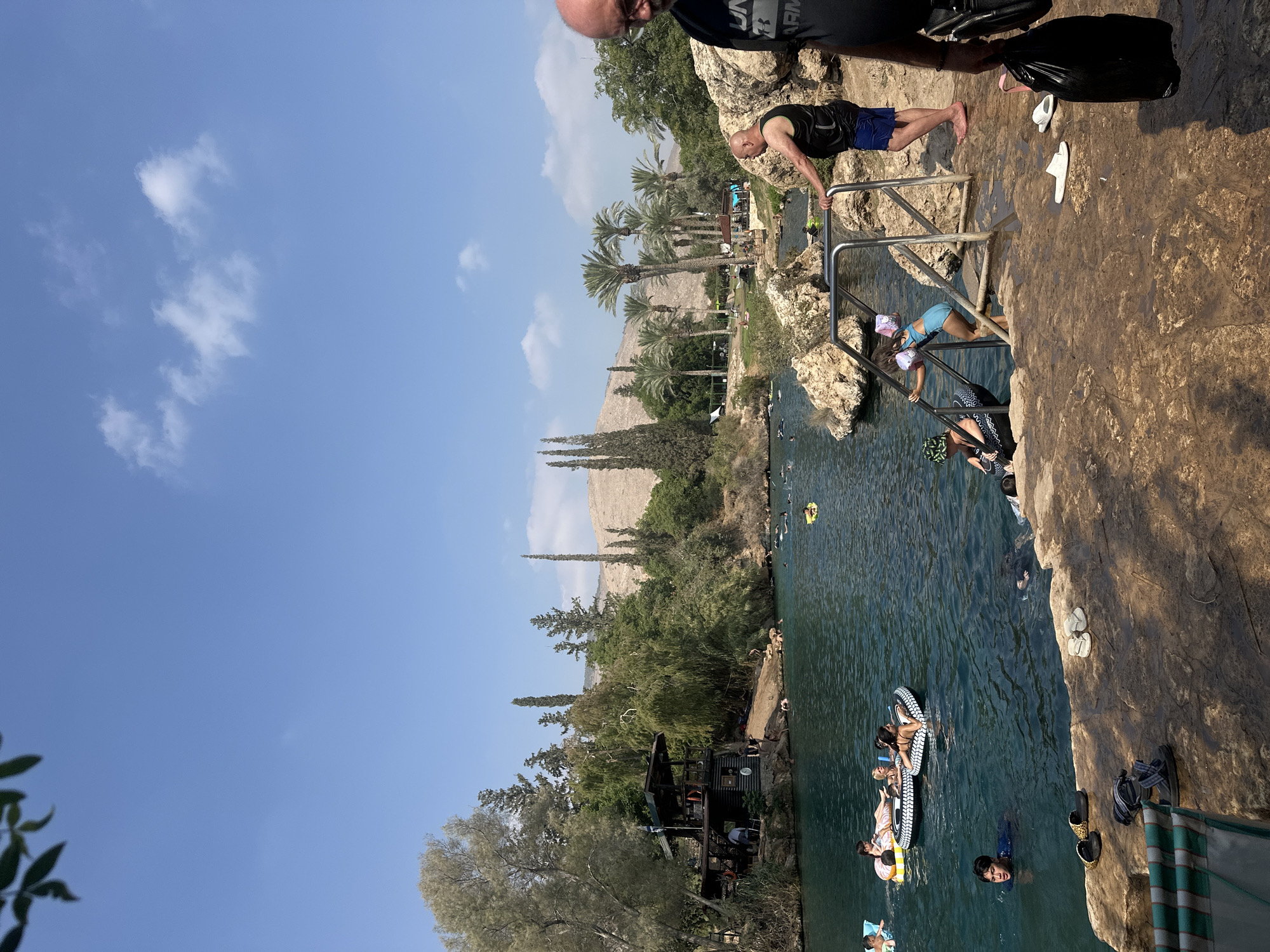 The image depicts a sunny day at a natural swimming area in Emek HaMaayanot Regional Council, Israel. The clear, blue water is populated with various individuals swimming and floating on inflatable rings. On the left side of the water, a two-story wooden structure with a balcony overlooks the scene, with people visible on its upper level. Lush green trees and palm trees line the banks of the water, and a rocky hillside is visible in the background. In the foreground, a rocky ledge is present, with several pairs of flip-flops and sandals scattered on its surface. A metal railing leads down to the water from this ledge, and a young girl in a blue swimsuit is ascending it. A man in a black tank top and blue shorts stands near the railing, looking out at the water. Further to the right, another man, partially visible, holds a black bag. The sky is a clear blue with a few scattered white clouds.
