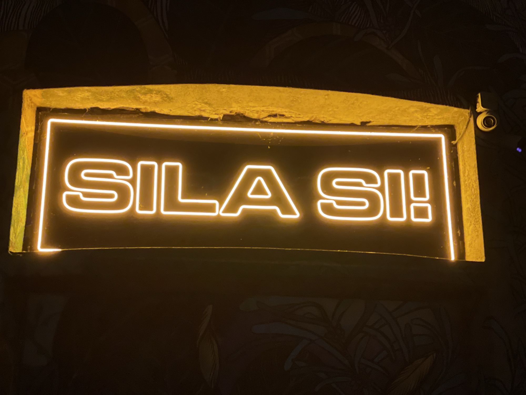 A brightly lit, rectangular sign displays the yellow neon text "SILA SI!" framed by a yellow border, and is mounted on a patterned wall of dark hues. A security camera is visible in the upper right corner. The location is Belgrade, Serbia.