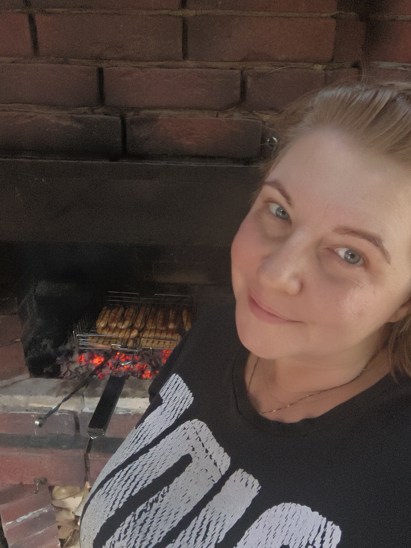 A medium shot photograph, taken indoors, shows a woman standing in front of a brick fireplace where hot coals are glowing beneath a grill filled with sausages. The woman, with light-colored hair pulled back and blue eyes, is wearing a black t-shirt with a white graphic print. She is looking towards the camera, with her right cheekbone prominently visible. The background consists of a dark, brick-lined fireplace. The overall scene suggests a casual outdoor cooking environment, possibly in the city of Vostets SNT, Russia.