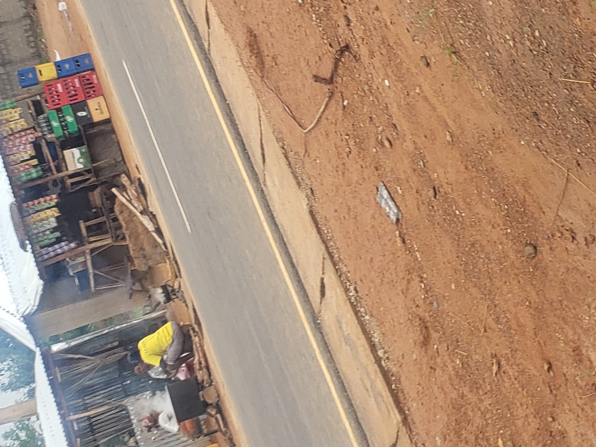 In Gumel, Nigeria, an outdoor food stall is situated next to a paved road with a yellow dividing line. Several individuals are present, with two actively engaged in preparing food. One person, wearing a bright yellow shirt and grey trousers, is crouched near a steaming pot. Another person, partially obscured, is also near the cooking area. The stall itself is constructed from corrugated metal and wood, displaying various goods on shelves, including rows of bottled beverages in colorful crates. The surrounding environment is earthy and arid, with scattered debris on the ground.