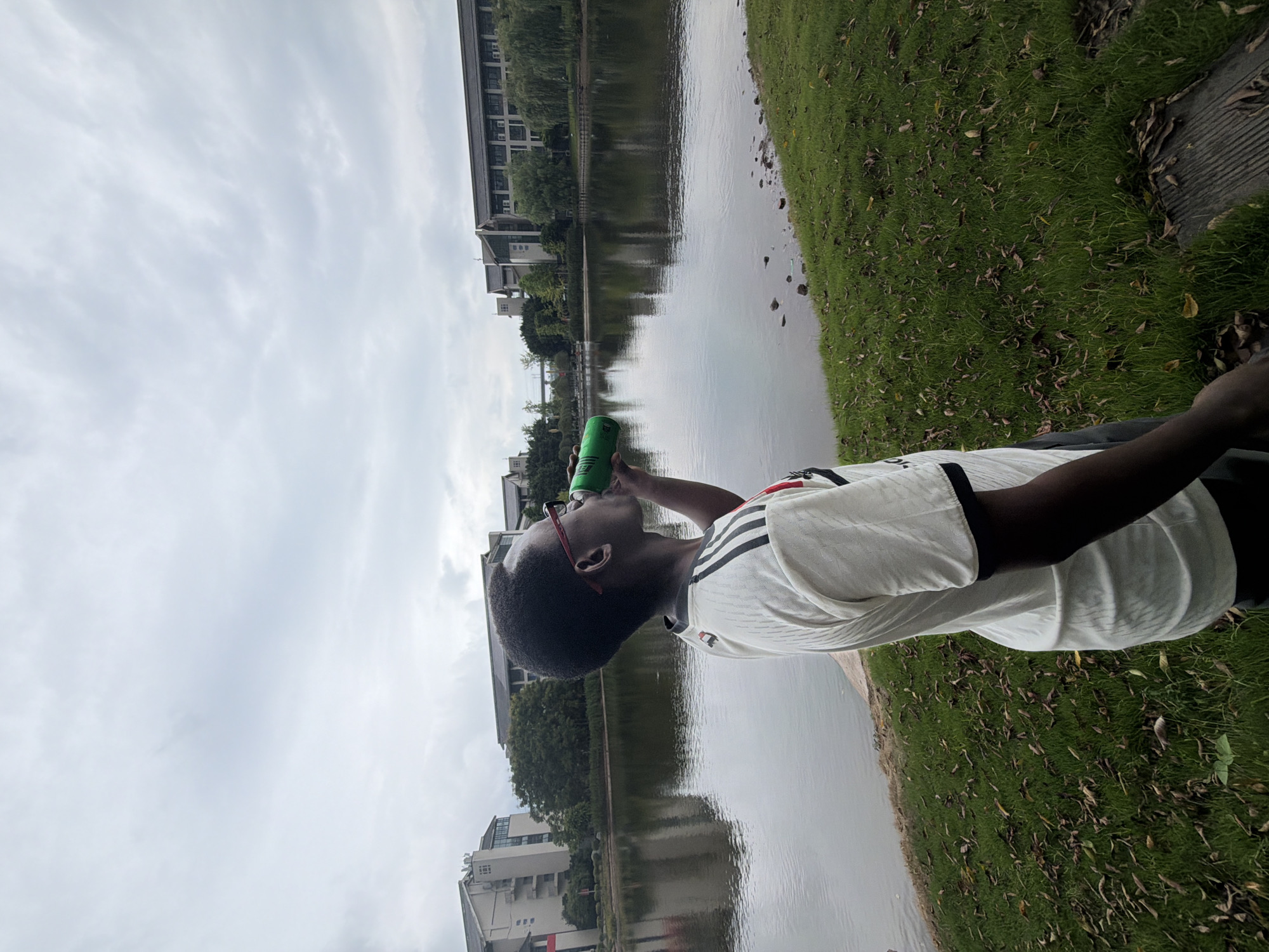 In Jiangjin, China, a young person with dark skin and red-rimmed glasses is standing on a grassy embankment, facing right and drinking from a green can. They are wearing a white t-shirt with black stripes on the sleeves and a red collar. Behind them, a tranquil lake reflects the overcast sky and the surrounding modern buildings, interspersed with trees. The grass is green but scattered with dry leaves, suggesting the season may be autumn. The scene appears to be a park or campus setting by a body of water.