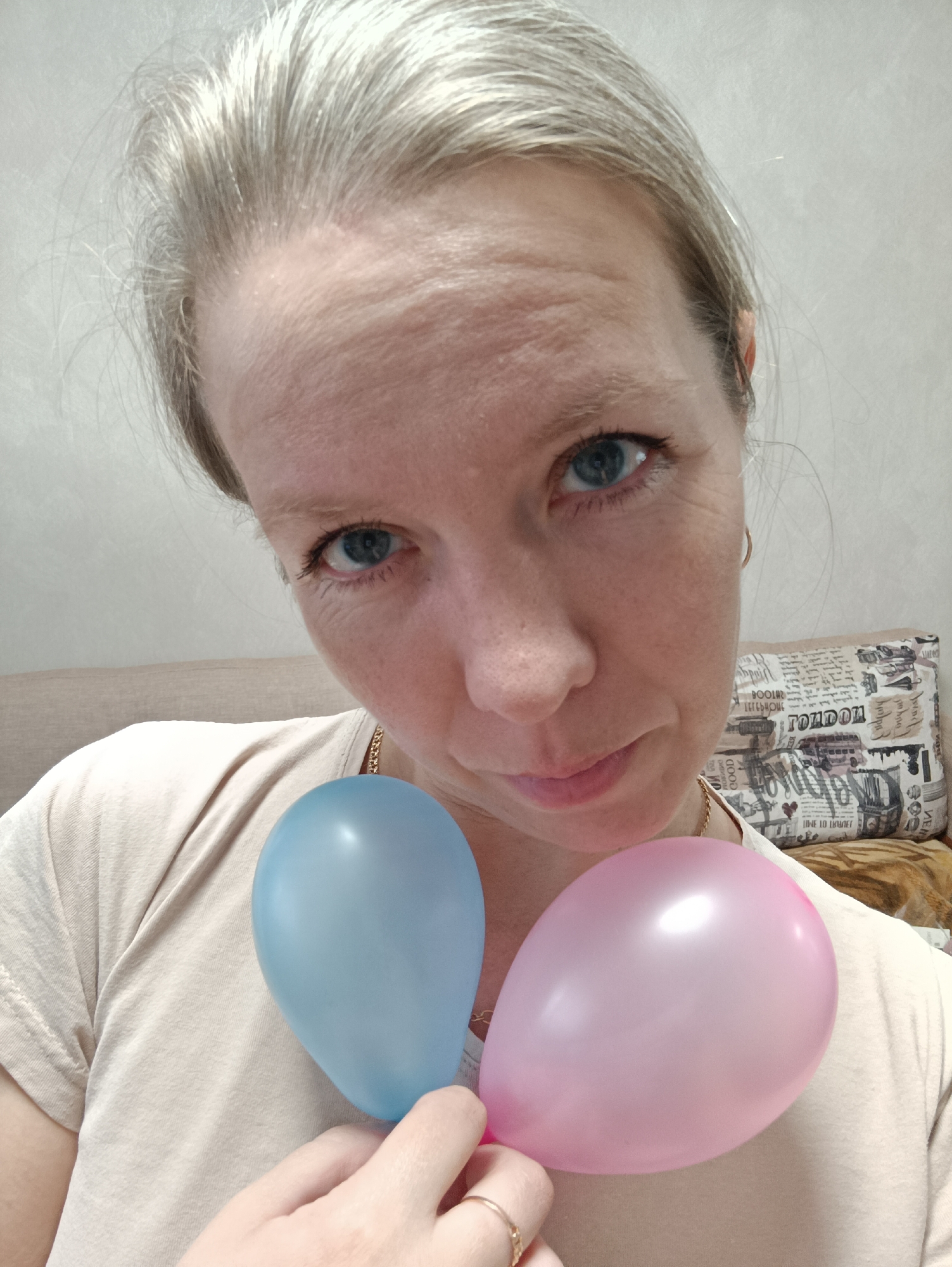 An adult woman is captured from the chest up, looking directly at the camera with a neutral to slightly contemplative expression. She has fair skin, blue eyes, and light blonde or graying hair pulled back from her face, revealing wrinkles on her forehead and around her eyes. She is wearing a plain, light-colored (beige/cream) t-shirt, and a thin gold chain is visible around her neck. On her left-hand ring finger, a gold ring is visible.
She holds two small, inflated balloons in her left hand, positioned over her chest. One balloon is light blue, and the other is light pink, typically symbolizing genders.
The background shows an indoor setting. To the left, a portion of a beige upholstered sofa or headboard is visible. To the right, a decorative pillow with a black and white newspaper-like print and illustrations is propped up. Clearly visible text on the pillow includes "LONDON" and "GOOD," along with graphics resembling a double-decker bus. The wall behind her is a plain, light color. The lighting suggests it is daytime. The image depicts a personal moment, possibly a gender reveal, within a home in Chaikovsky, Russia.