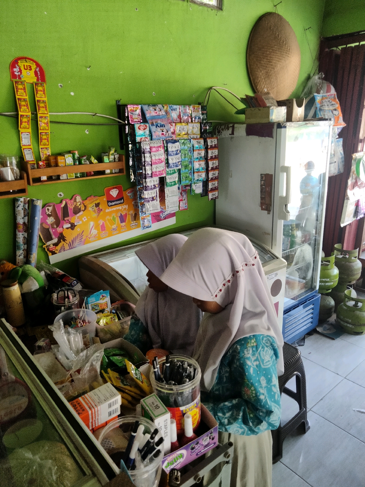 This is a photograph taken inside a small shop in Sidorejo, Indonesia. The shop is filled with various goods, suggesting it functions as a convenience store or warung. Two young girls, both wearing headscarves and modest clothing, are the main subjects. One girl, on the left, is partially obscured, but her headscarf is visible. The other girl, on the right, is more fully visible, wearing a white headscarf and a teal and white patterned tunic. They appear to be standing behind a counter laden with products.
The environment is rustic. The walls are painted a vibrant green. Shelves on the wall display an assortment of packaged items, including what appear to be snacks, toiletries, and medicines. A prominent display of "U3" branded products hangs on the left wall, alongside other items on a wooden shelf. A large, light-colored refrigerator stands against the back wall, its glass door reflecting the interior. Various items are piled on top of and around the refrigerator, including what looks like a woven conical hat, bags of goods, and a blue plastic crate.
On the counter in front of the girls, there are numerous items, including plastic containers filled with small items, possibly candy or snacks, packaged food items, and jars filled with pens or pencils. A large poster depicting ice cream with the brand "Diamond" is displayed on the wall behind the counter. Several green gas cylinders are visible outside the shop to the right. The floor is tiled with a gray and white pattern. The lighting suggests it is daytime, and the overall impression is of a busy, functional, small-scale retail space.