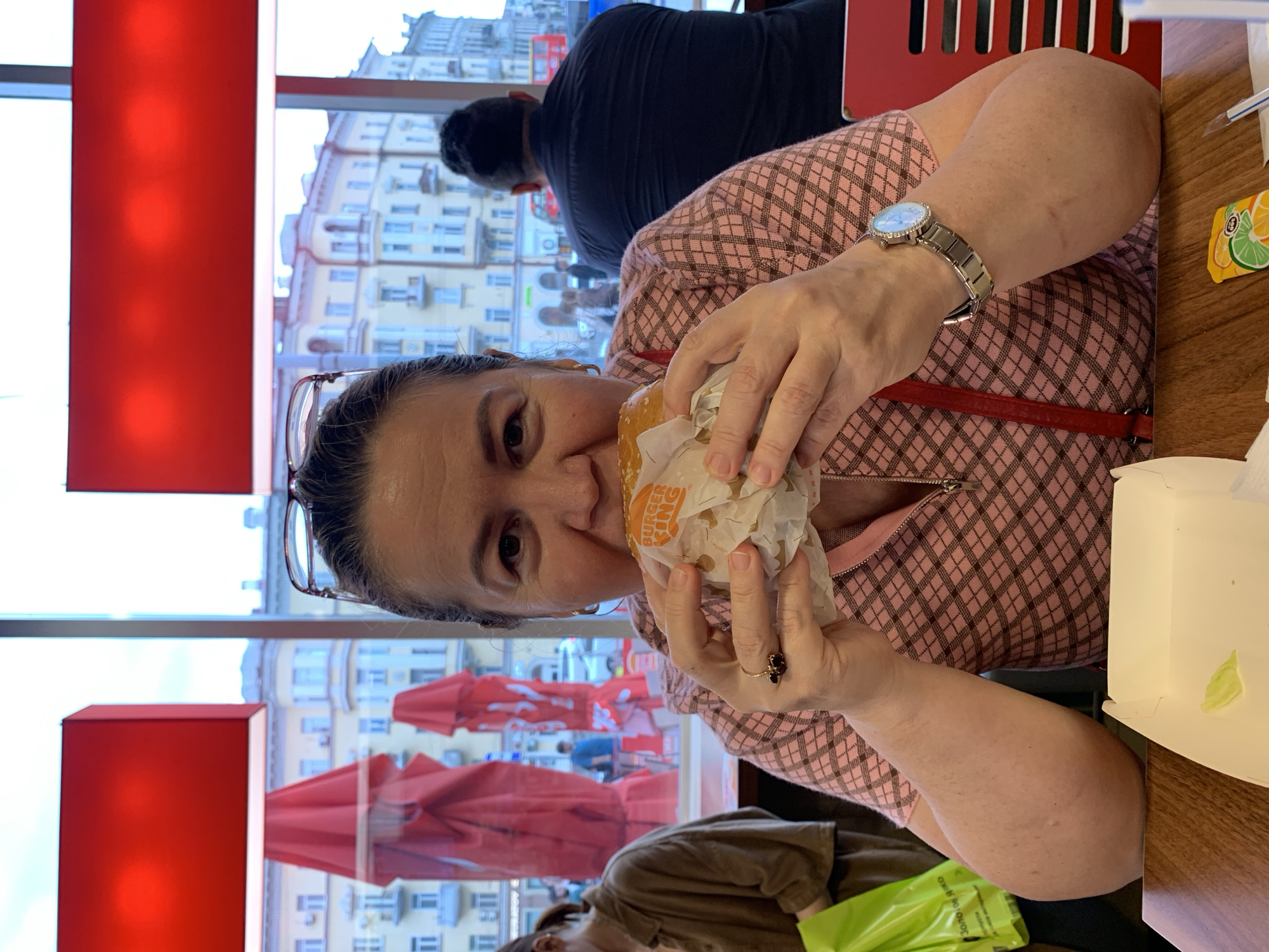 A middle-aged woman with dark hair and glasses is eating a Burger King hamburger. She is wearing a pink and brown checkered shirt with a zipper. She has a ring on her left hand and a watch on her right wrist. The woman is seated at a wooden table, in front of a large window. Outside the window, a street scene in Kazan, Russia, can be seen. Buildings with classical architecture and red umbrellas are visible. A man with dark hair is sitting behind the woman, facing away from the camera. A green plastic bag is on the left side of the table. A white takeout box is also on the table.