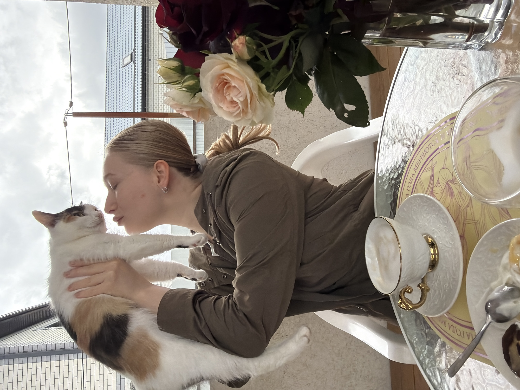 A young woman with blonde hair pulled back in a ponytail is holding a calico cat in her arms and appears to be kissing its nose. The woman is wearing a green jacket and has a small hoop earring. The cat is predominantly white with patches of black, tan, and brown. They are positioned next to a table with a glass top, on which there is a vase of red and white roses, a teacup and saucer, a plate with a spoon, and a glass bowl containing a clear liquid. The table sits on a white plastic chair. The background shows a building with a gray tiled roof and utility poles against a cloudy sky. The location is Rossiyskiy, Russia.
