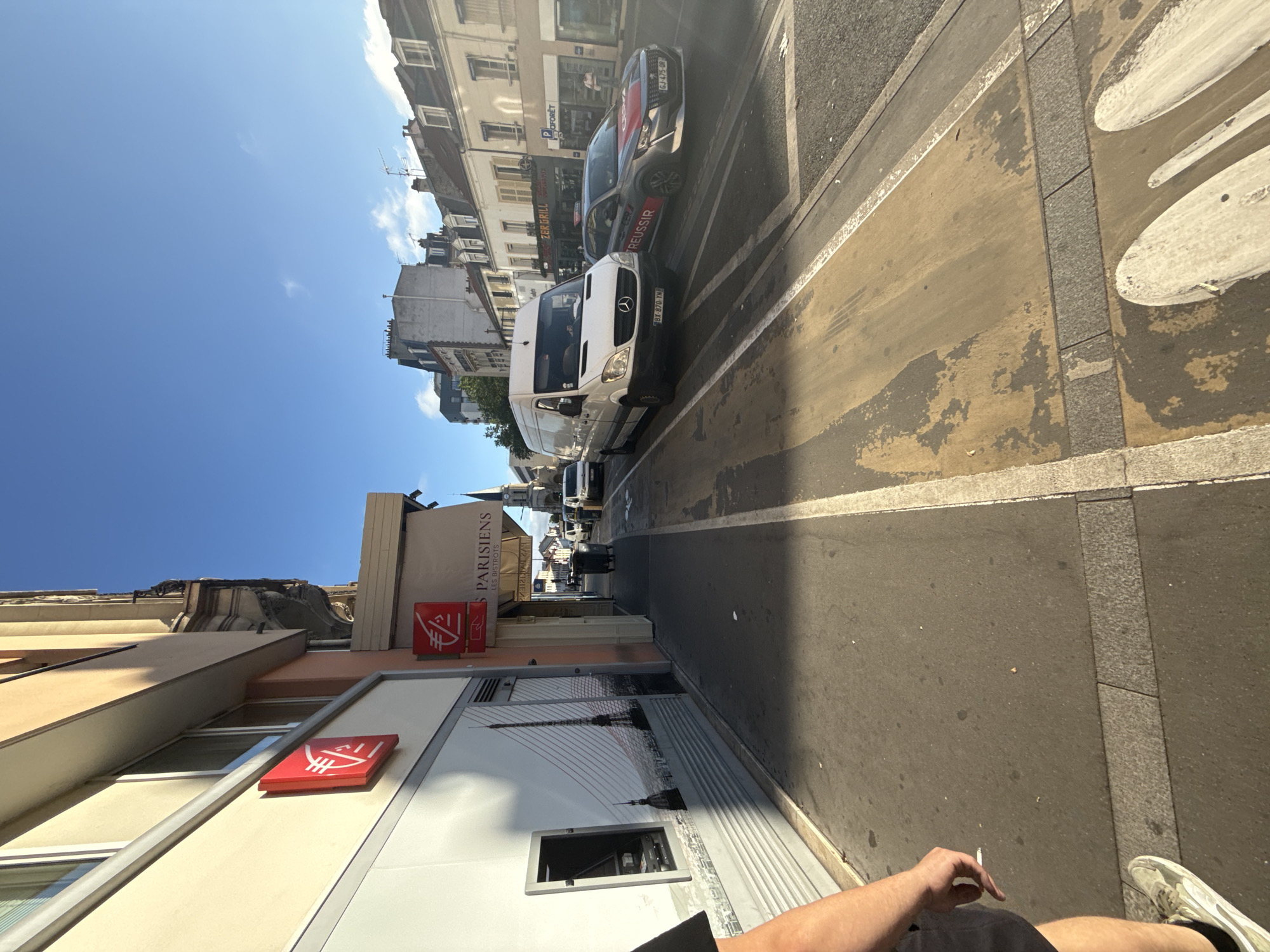 The image depicts a street scene in Colombes, France, under a clear blue sky. On the left, a building facade features a red square sign with a white logo, likely representing a bank, and a large mural of the Eiffel Tower. A person's legs and arm are visible in the foreground on the left, holding a cigarette.
The street itself has a marked lane for vehicles and a pedestrian walkway. Several vehicles are present: a white van with a Mercedes-Benz logo, a dark gray car with red and white decals, and other cars further down the street. Buildings line the right side of the street, with some displaying signs for businesses such as "ZER GRILL" and "FORÊT". In the background, a church spire is visible. The environment appears to be an urban setting, possibly a commercial or residential street. The lighting suggests it is daytime, with sunlight casting shadows.