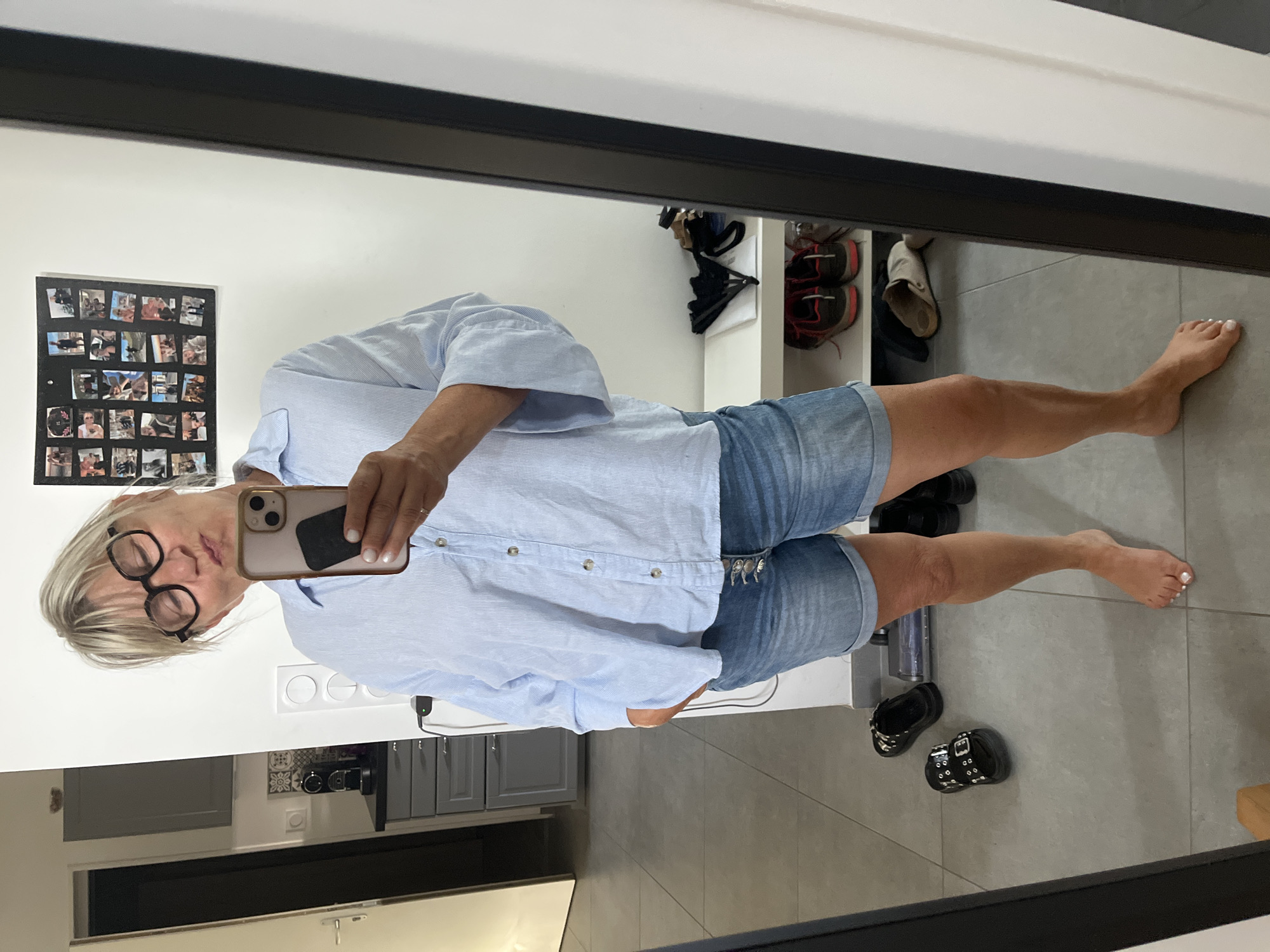 A woman is taking a mirror selfie in what appears to be a hallway or entryway of a home. She is wearing a light blue striped button-down shirt, denim shorts, and black sandals. She has blonde hair and is wearing large-rimmed glasses. Her lips are pursed as if she is blowing a kiss. In the background, there is a collage of photos on the wall, a doorway leading to another room, and a shoe rack with several pairs of shoes. The floor is tiled. The woman's left foot is bare. It is not possible to determine the exact location within Chambœuf, France from the visual information provided.