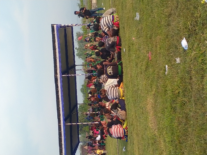 The image depicts an outdoor gathering on a grassy field in Glonggong, Indonesia. In the foreground, green grass contains scattered litter, including plastic bags and white debris. A group of approximately 10-15 individuals are seated in a semi-circle on the grass. Several wear horizontally striped shirts, while others wear dark shirts, one with a gold graphic (possibly "BLACK MANGUS") and another with a red sash. Some seated individuals have visible sashes or belts in yellow, green, or red. In the midground, a large temporary tent structure with a blue and white striped canopy is erected. Beneath and around this structure, a larger crowd of people is gathered, some seated on green plastic chairs, others standing. To the far right, a male individual stands, operating a video camera mounted on a tripod, directed towards the main gathering. The background features trees, distant structures, and a mountain range under a pale blue, slightly hazy sky. The scene is illuminated by natural daylight.