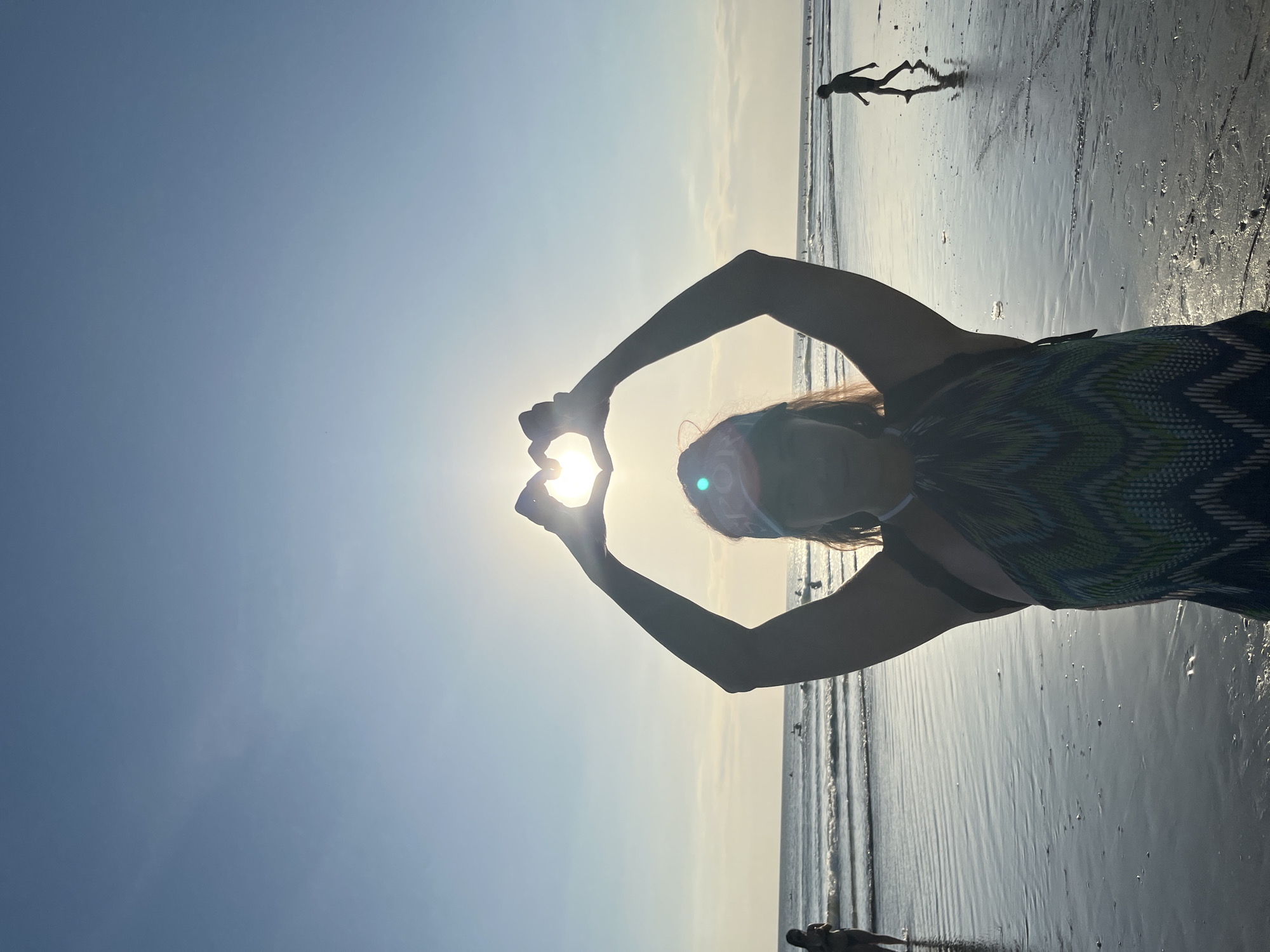 The image depicts a person on a beach at sunset, with the sun positioned to form a heart shape between their hands. The setting is Kuta, Indonesia. The sky is a gradient of blue and yellow, indicating it's late afternoon or early evening. The sand is wet, reflecting the light of the setting sun, and small waves are visible in the background. Another person is silhouetted further down the beach, appearing to be walking or running. The overall mood is peaceful and serene, with a sense of joy captured by the heart gesture. The person in the foreground is wearing a patterned top and a head covering. No visible text is present in the image.