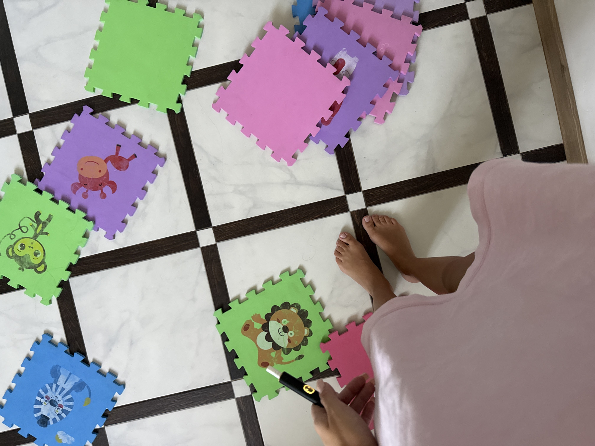 The image shows a floor with interlocking foam play mats. Some of the mats are plain colored (blue, green, pink, purple) while others have cartoon animal images printed on them (a tiger, a monkey, a giraffe, a lion). A person's bare feet and lower legs are visible in the bottom right of the image. One of the person's hands is in the upper left, holding a black electronic device with a yellow light. The floor appears to be tiled with a marble-like pattern and dark brown grout lines, suggesting an indoor setting. There are no landmarks or indications that would place this scene specifically in Chișinău, Moldova.