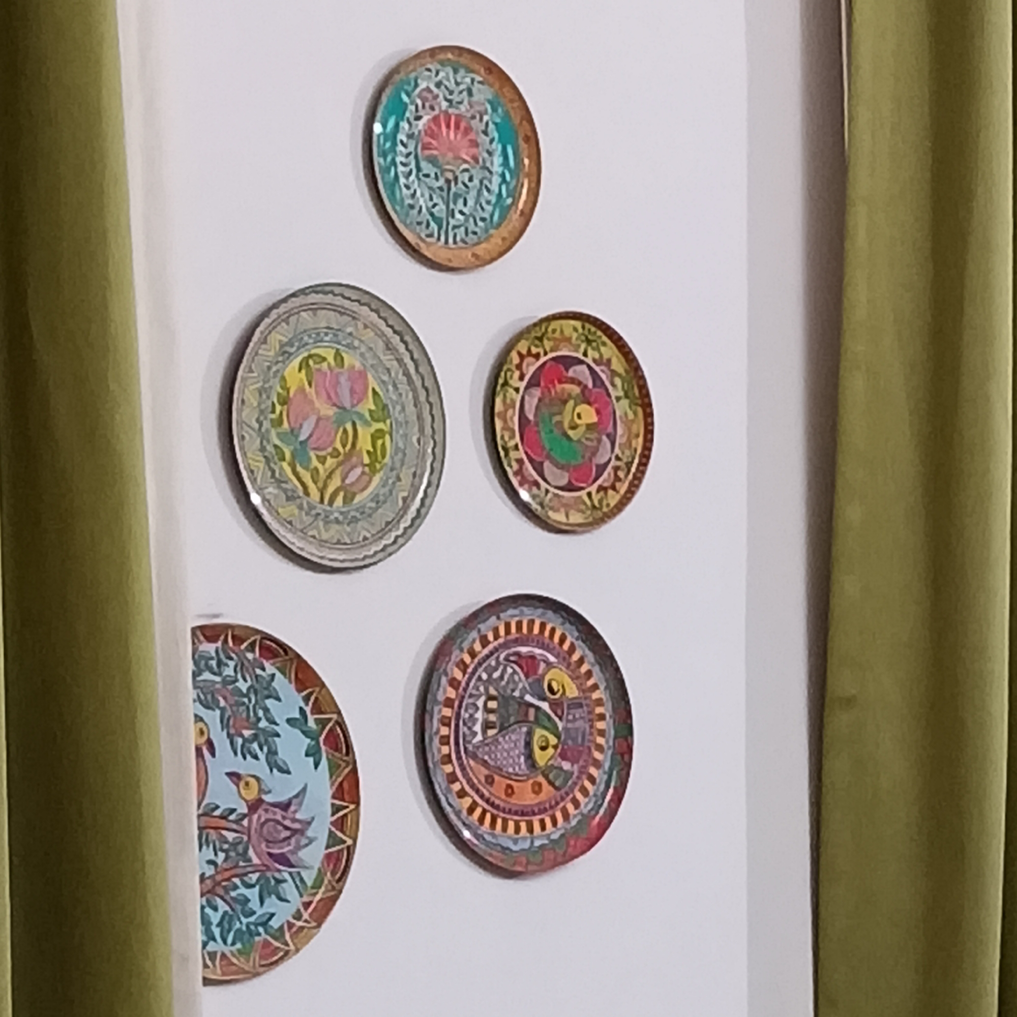 The image shows five decorative plates hanging on a white wall. The plates vary in design, with floral and bird motifs. A green curtain partially frames the right side of the image and a similarly colored object the left side of the image. No people are visible. The location is identified as Calangute, India, although the image does not provide direct visual evidence of this location beyond the style of the decorative plates.