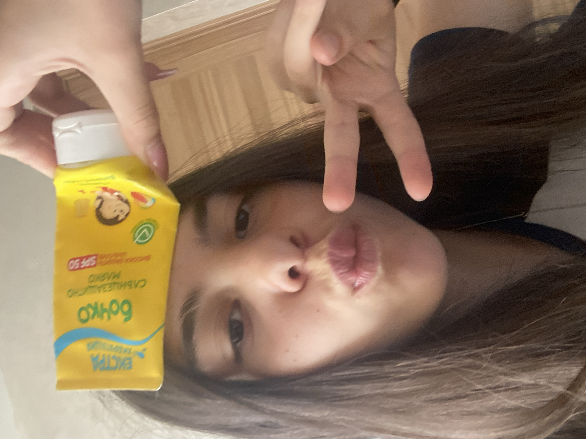 A young woman is pictured holding a yellow bottle labeled "BONKO Слънцезащитно мляко SPF 50 ЕКСТРА." She is making a peace sign with her other hand and pursing her lips. Her hair is dark brown and wavy. The background is out of focus and appears to be an interior space with wooden features. There is no evidence to suggest that the location is Dragovishtitsa, Bulgaria.