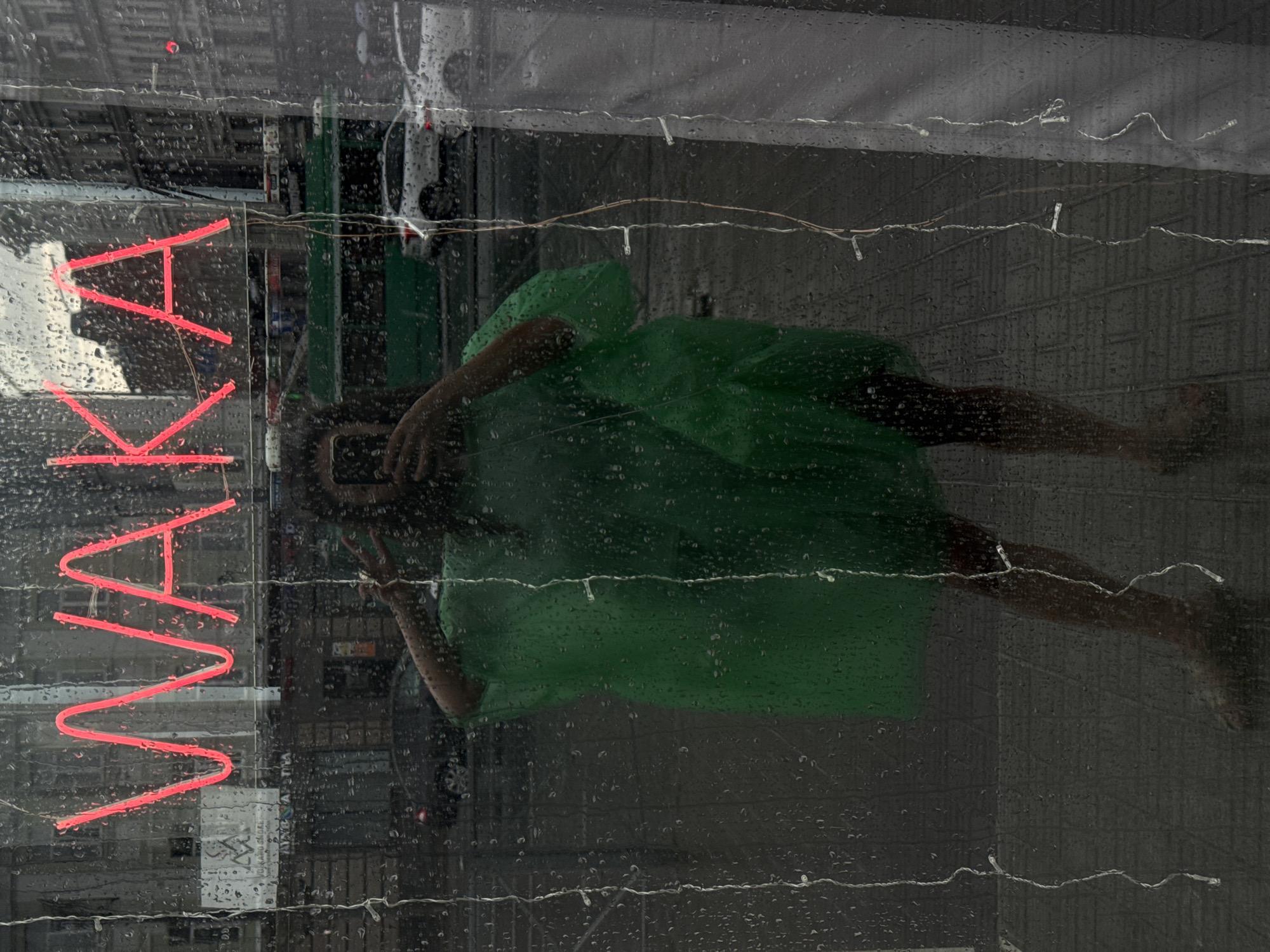 The image, taken through a rain-streaked window in Kazan, Russia, shows a person wearing a green poncho, holding a smartphone to take a selfie. The neon sign "WAKA" glows in red behind them. The reflection in the window reveals a street scene with buildings, a green tram, and cars. The ground is wet from the rain, and the overall atmosphere is damp and overcast.