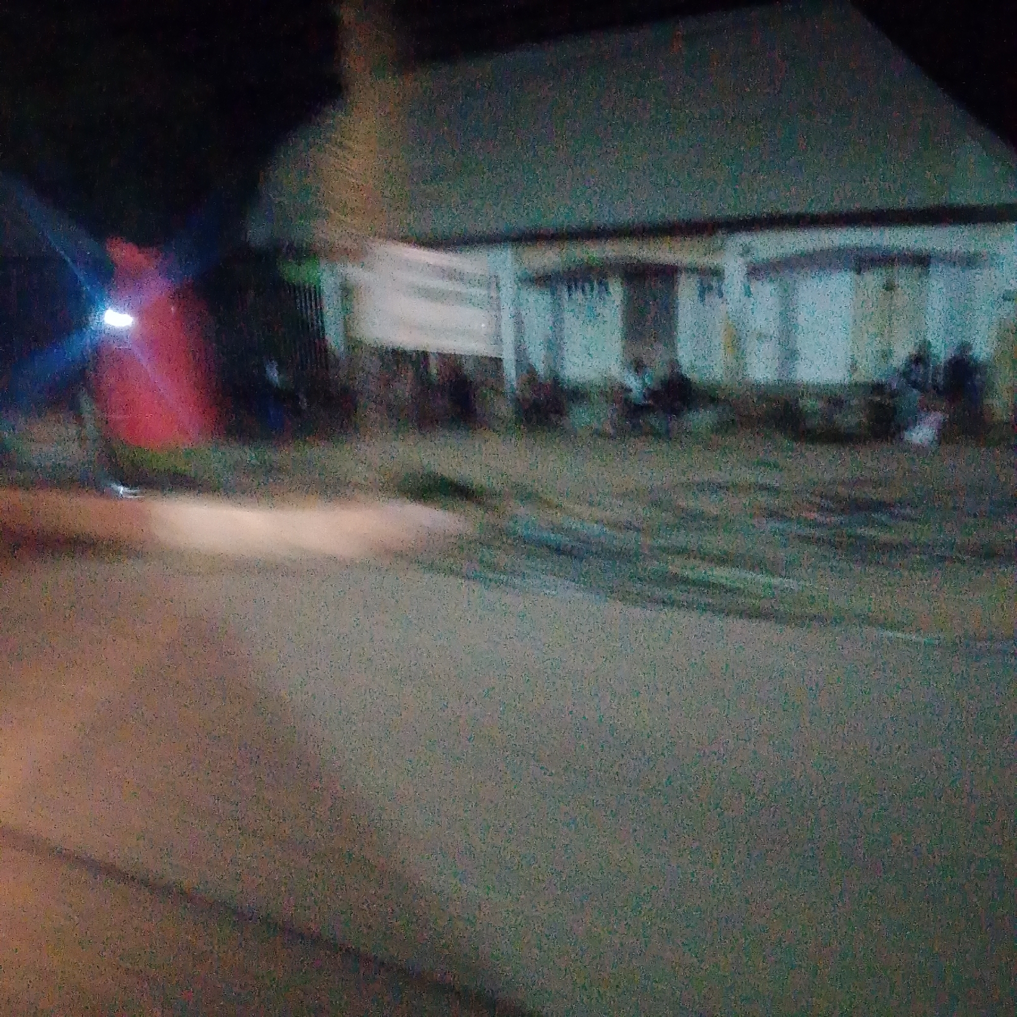 The image depicts a nighttime scene in the city of Jalingo, Nigeria. In the background, a building with a light-colored facade and a dark roof is visible. A banner or sign with white and some blue lettering is draped across the front of the building. The building appears to be a commercial establishment, possibly a post office, given the visible letters "POR" on the sign.
In front of the building, several figures are blurred due to the low light conditions and motion. They appear to be seated or standing, suggesting a gathering of people. To the left of the building, a bright red object is illuminated, possibly a vehicle or a structure emitting light. A strong blue light source is also present near this red object.
The foreground shows a paved surface, likely a road or a sidewalk, with some blurry white light streaks, indicating movement or reflections from light sources. The overall ambiance is that of a dimly lit street scene at night.