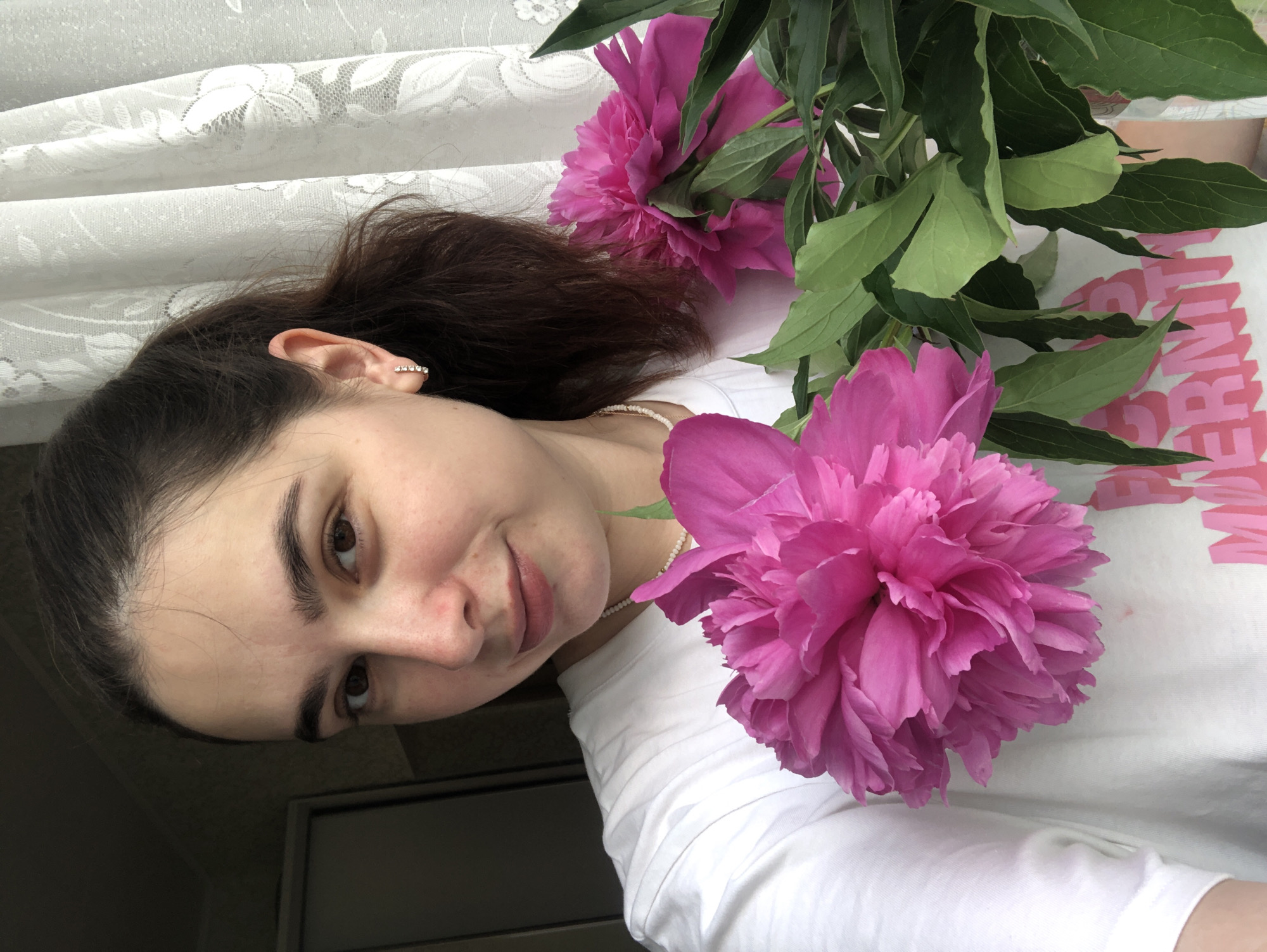 In an indoor setting, likely a home, a young woman with dark hair pulled back in a ponytail poses for a selfie. She is wearing a white t-shirt and a pearl necklace. She has a small, stud earring in her left ear. She is holding a bouquet of vibrant pink peonies, with their large, layered petals and green leaves prominently displayed. The background features a white lace curtain and a partially visible dark doorway on the left. The lighting suggests it might be daytime, with natural light filtering in from the right. The specific location in Noviy, Russia, cannot be determined from the visual information provided.