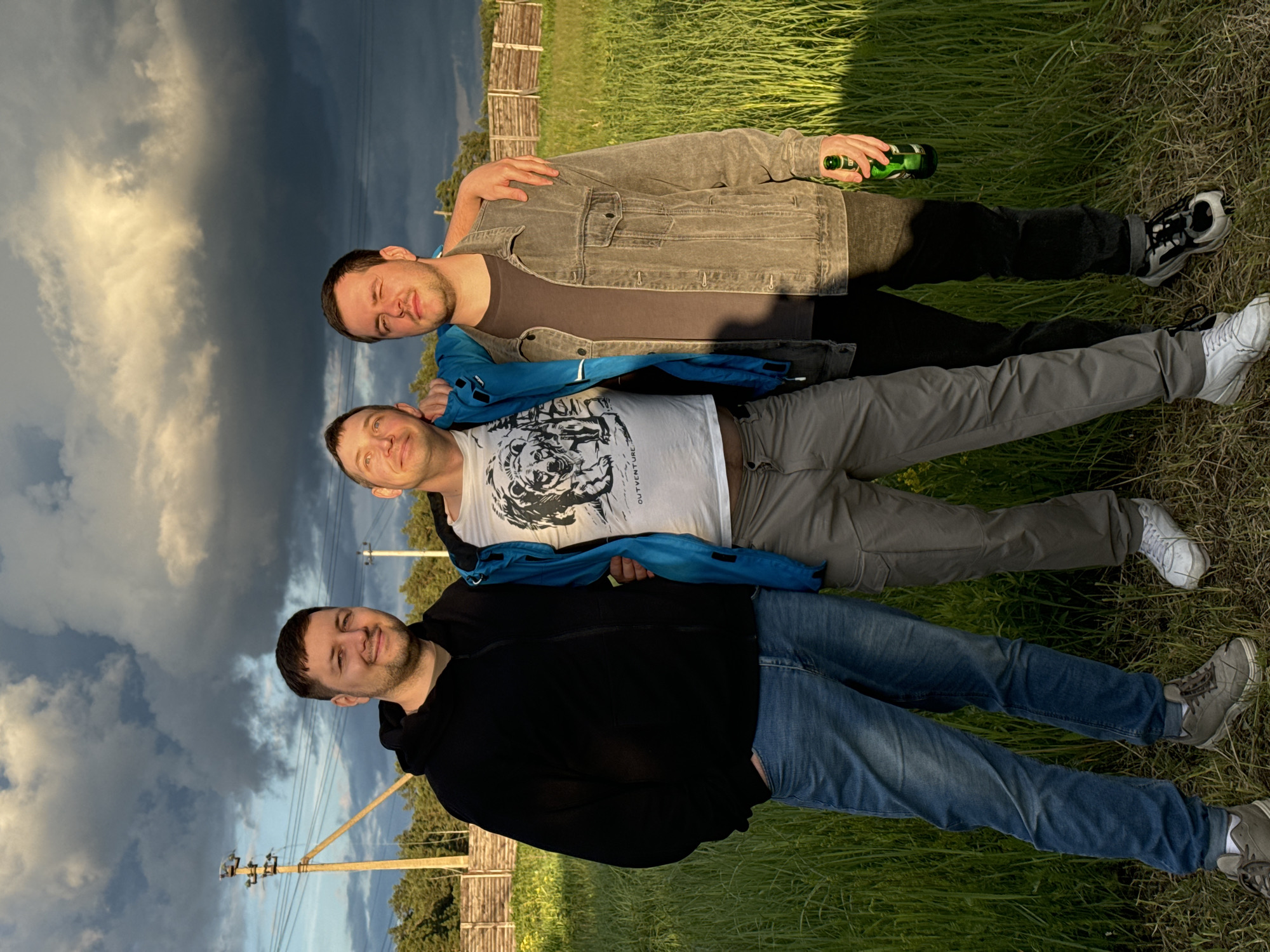 Three men are lying on the grass in Vysokinichi, Russia, facing the camera. The man at the top is holding a green bottle and has his arm around the man below him. All three men are fully clothed. The background includes a fence, electrical poles, and a partly cloudy sky.