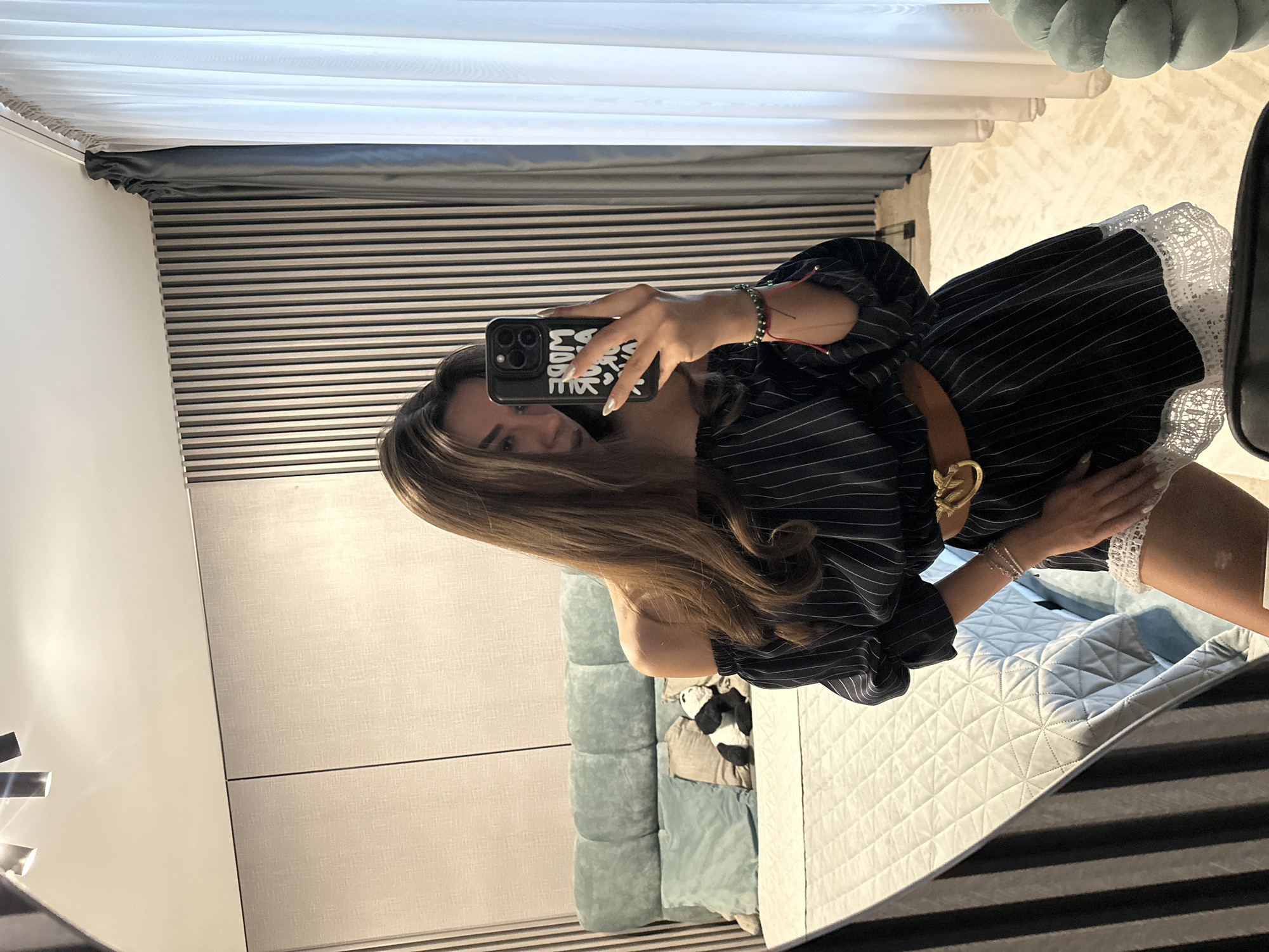 A young woman with long, brown hair is taking a mirror selfie in what appears to be a bedroom. She is wearing an off-the-shoulder, pinstriped dress with white lace trim at the hem, a brown belt with a gold buckle, and several bracelets. She is holding a black smartphone with a white design on the back. Behind her, a bed with a light blue headboard and quilted bedding is visible. A stuffed panda bear rests on the bed. The room features a wall with vertical wooden slats and light-colored paneling, and a sheer white curtain is visible on the right side of the image. The floor is covered with a light-colored rug. The location is not specified in the image or its metadata.