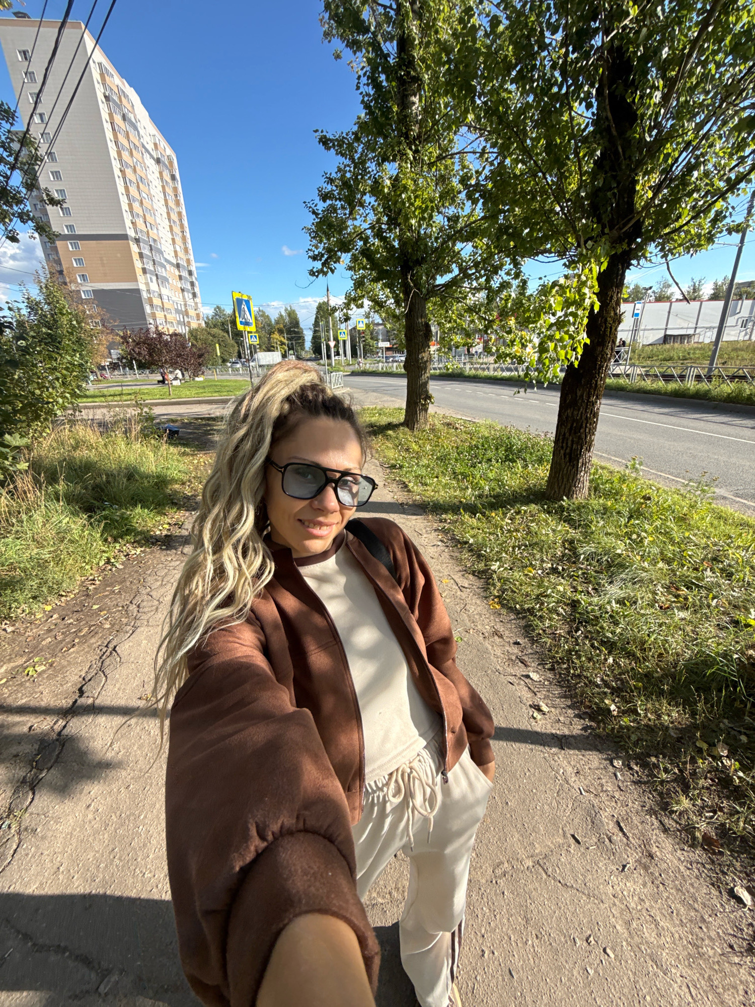 In a sunny outdoor setting, a young woman with blonde, braided hair pulled into a high ponytail and large dark sunglasses stands on a cracked asphalt pathway. She is wearing a brown zip-up jacket over a cream-colored t-shirt and matching cream-colored drawstring pants. Her left hand is in her pocket, and her right arm is extended, holding a phone to take a selfie.
Behind her, the pathway is bordered by dry grass and fallen leaves. Further back, a paved road runs parallel to the pathway, with trees lining the edge. In the background, a tall, modern apartment building with a beige and grey facade stands against a clear blue sky. Pedestrian crossing signs are visible along the road, and there are glimpses of other buildings and greenery in the distance. The overall impression is an urban environment on a bright, clear day. This scene is located in the city of Rusanovo, Russia.