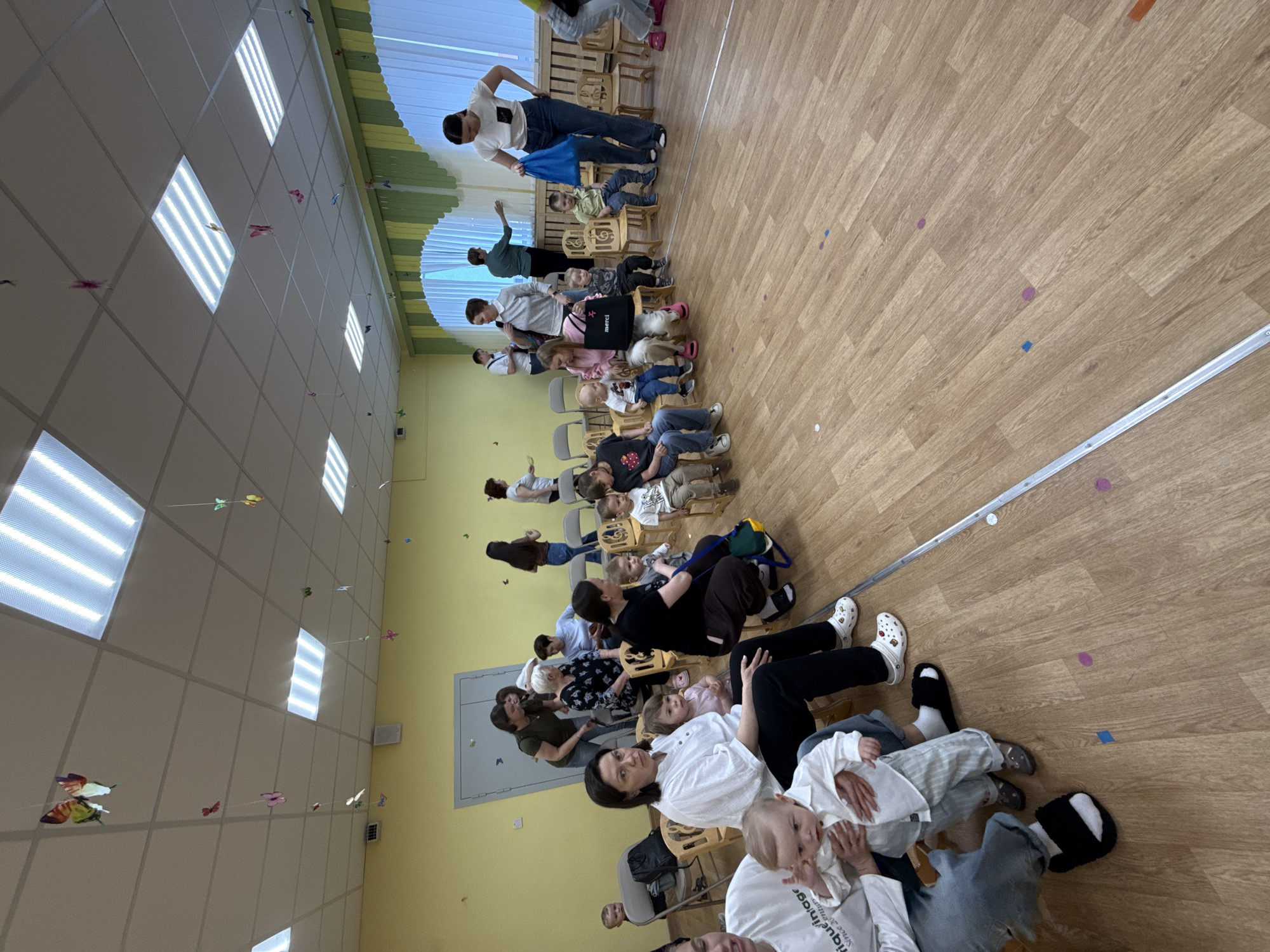 The image shows a group of adults and children sitting on chairs in a room with a wooden floor and yellow walls. The ceiling has fluorescent lights and is decorated with hanging butterflies. The people are arranged in rows, and some are holding young children. There appears to be an event or gathering taking place. The location is in the city of Шебунино, Russia.