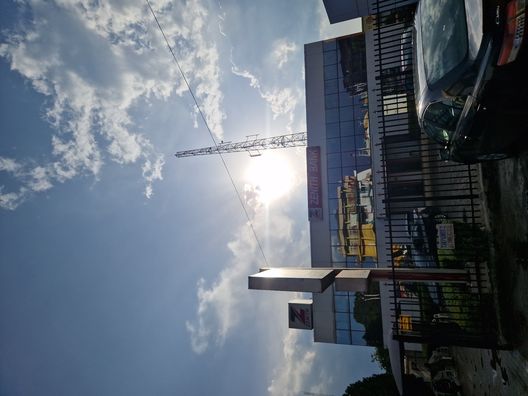 The image captures the Zenith Bank building in Jos, Nigeria, under a partially cloudy sky with the sun shining brightly. A prominent cell tower stands to the right of the bank building. The bank's facade features large, reflective glass windows, displaying a mirrored image of buildings across the street, including a yellow and beige structure. In front of the bank, a black sedan is parked, and a black metal fence encloses the property. A sign is visible on the fence, reading "CAR PARKED AT OWNER'S RISK." To the left of the bank, a smaller Zenith Bank sign is mounted on a pillar. The ground appears wet in places, suggesting recent rain. The overall impression is that of a typical urban commercial scene.