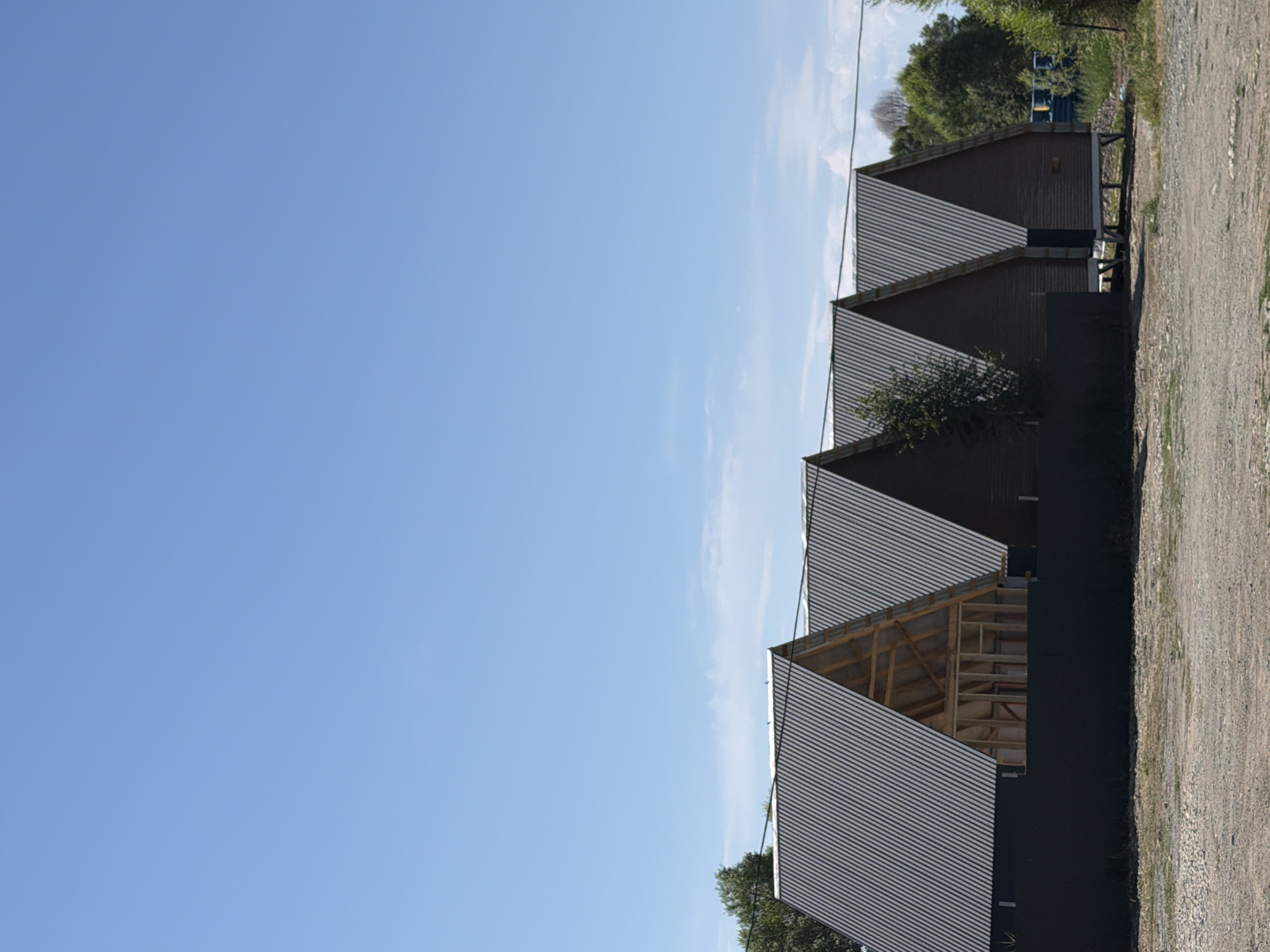 The image shows a row of modern, angular buildings with dark brown wooden siding and corrugated metal roofs. The buildings are arranged in a stepped pattern, receding into the distance. The closest building's roof is partially open, revealing the wooden structure underneath. The ground in front of the buildings is a gravel path, and a dark fence runs along the edge of the property. Behind the buildings, there are trees and a clear blue sky with some wispy clouds. The overall impression is one of contemporary architecture in a somewhat rural or developing setting. The time of day appears to be daytime, likely morning or afternoon, given the bright sunlight and clear sky. There are no people or animals visible in the image. No text is visible.