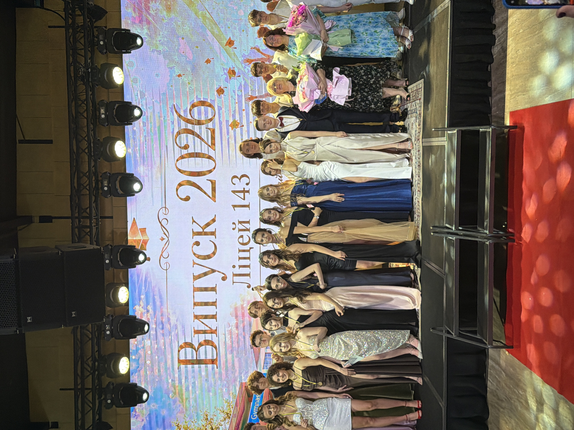 A large group of young adults, likely graduating students, pose for a photograph on a stage in front of a large digital screen displaying "Випуск 2026" (Graduation 2026) and "Ліцей 143" (Lyceum 143). The students are dressed in formal attire, with many of the young women wearing long, elegant dresses and the young men in suits. Some of the students are holding awards or bouquets of flowers. There are two women in the center holding large bouquets. The stage is illuminated by spotlights, and a red carpet is visible in the foreground. The background screen shows a graduation cap motif.