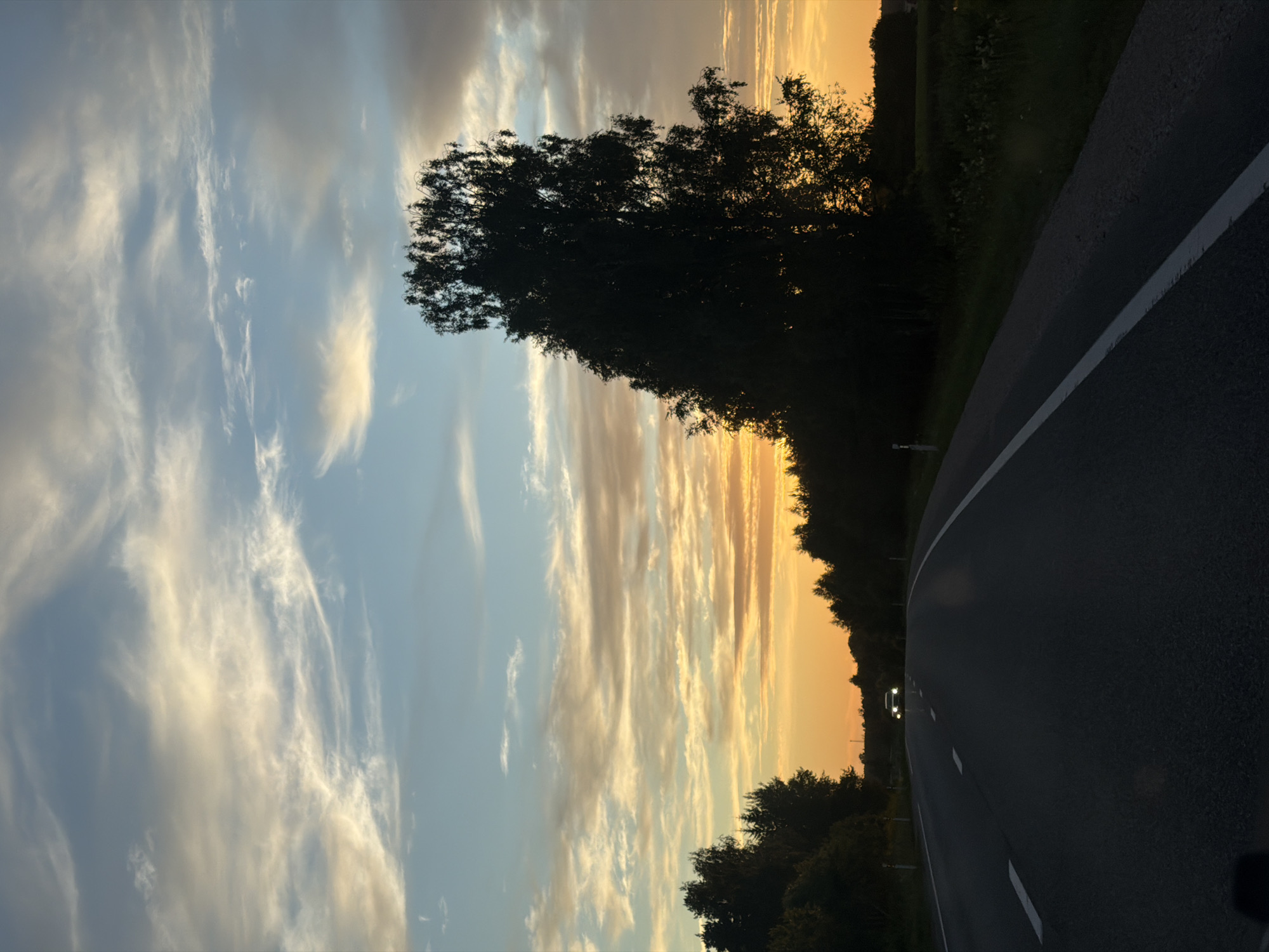 This image depicts a rural road in Aizupes, Latvia, during a sunset. The sky is filled with wispy clouds streaked with orange, yellow, and blue hues. A large, dark silhouette of a tree stands prominently on the right side of the frame. The road itself is asphalt with white lane markings and stretches into the distance, where a single car with its headlights on is approaching. The surrounding landscape is characterized by dark silhouettes of trees and vegetation, suggesting a wooded area or fields bordering the road. The overall atmosphere is serene and atmospheric, indicative of the twilight hours.
