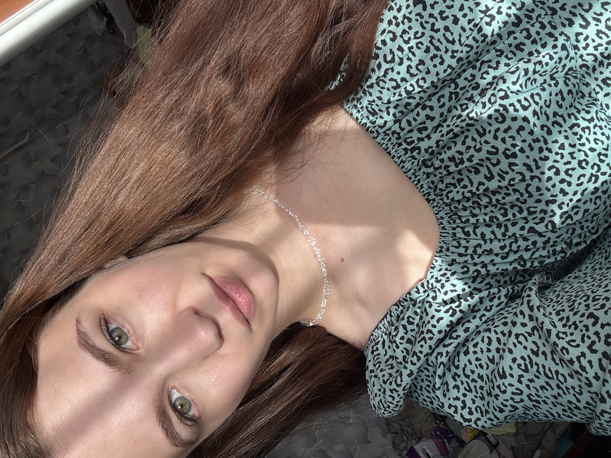 The image shows a close-up, self-portrait of a young woman with long, brown hair and green eyes. She is wearing a light blue, leopard-print off-the-shoulder top and a delicate, clear beaded necklace. A sunbeam crosses her forehead, casting a diagonal shadow. The background is blurred and appears to be an indoor setting with some patterned fabric and a hint of furniture. The woman is positioned in a way that her face and upper body are the primary focus. The scene is devoid of any explicit indicators of location within Nizhnyaya Gar', Russia.