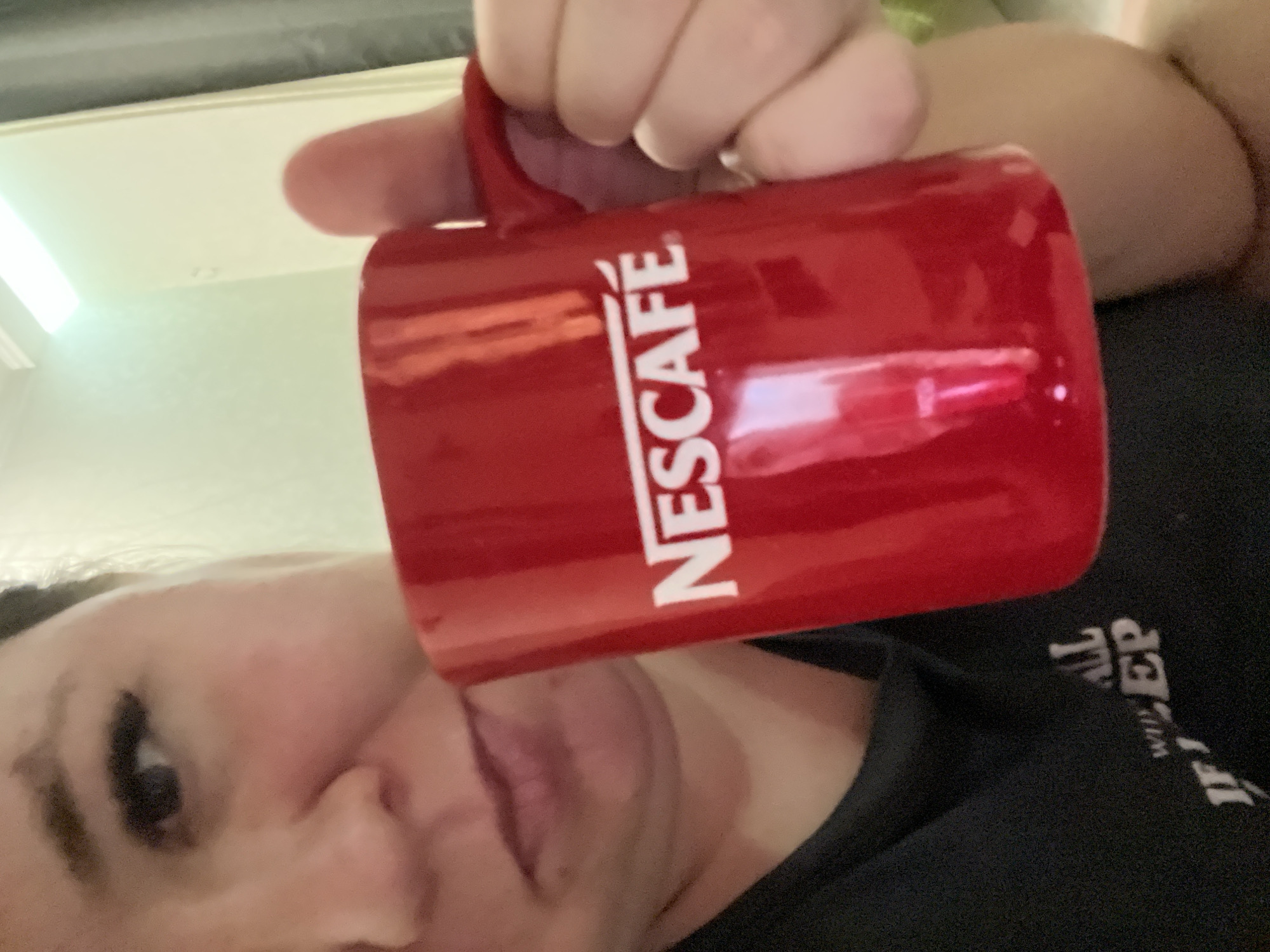 A person holds a red Nescafé mug. The person wears a black shirt with white writing. The location is potentially a room in Chaikovsky, Russia, given the Nescafé brand's availability there. More details are needed for a definitive identification.